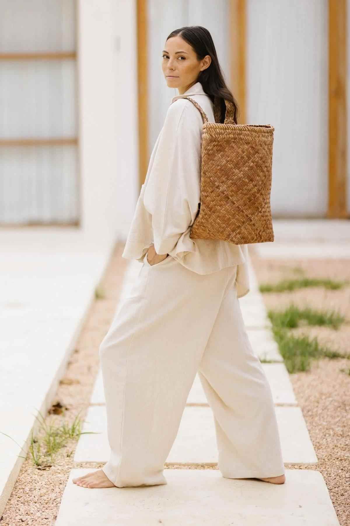 A person with long dark hair stands barefoot on a pathway, clad in the Grace Jacket & Mona Pant Set by Myrah Penaloza. The light-colored 100% cotton linen ensemble complements their look as they carry a large, woven backpack and glance over their shoulder at the camera. The background features a building with wooden frames and white walls.