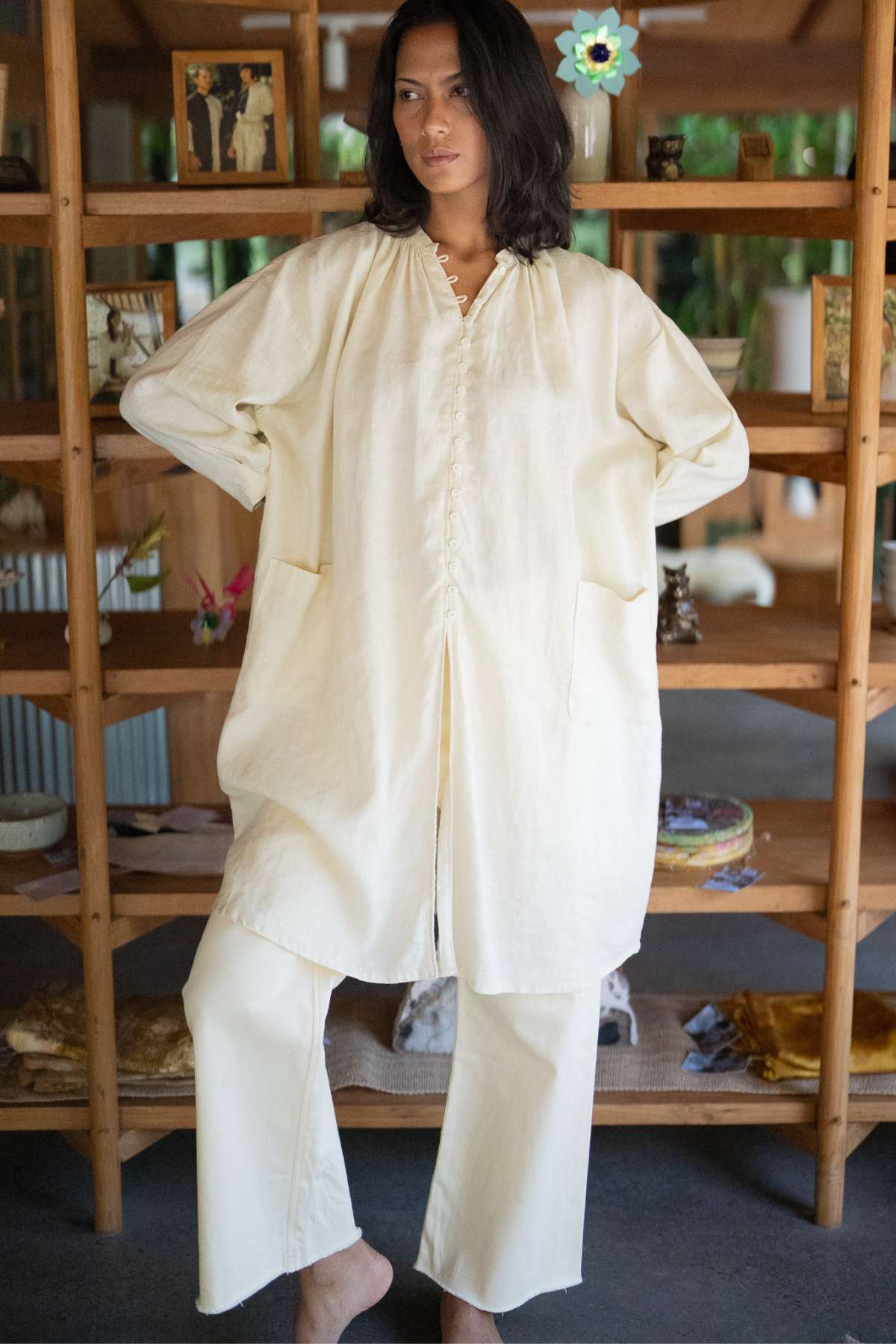 Dressed in a cream-colored ensemble, an individual exudes calm relaxation while standing in a room adorned with wooden shelves displaying various small objects and decor items. They are wearing a long Zazen Linen Top from Myrah Penaloza, paired with matching pants for an effortlessly elegant look.