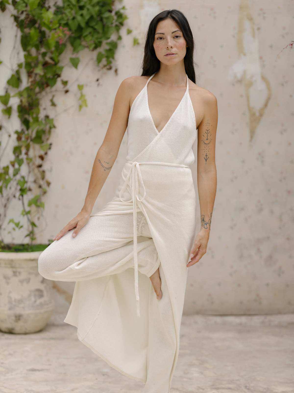 A woman with long dark hair stands in a Myrah Penaloza Honey Cotton Knit Wrap Dress, posing in a yoga tree pose. Her arms are relaxed, showcasing her tattoos, and the background features climbing plants against a light-colored wall.