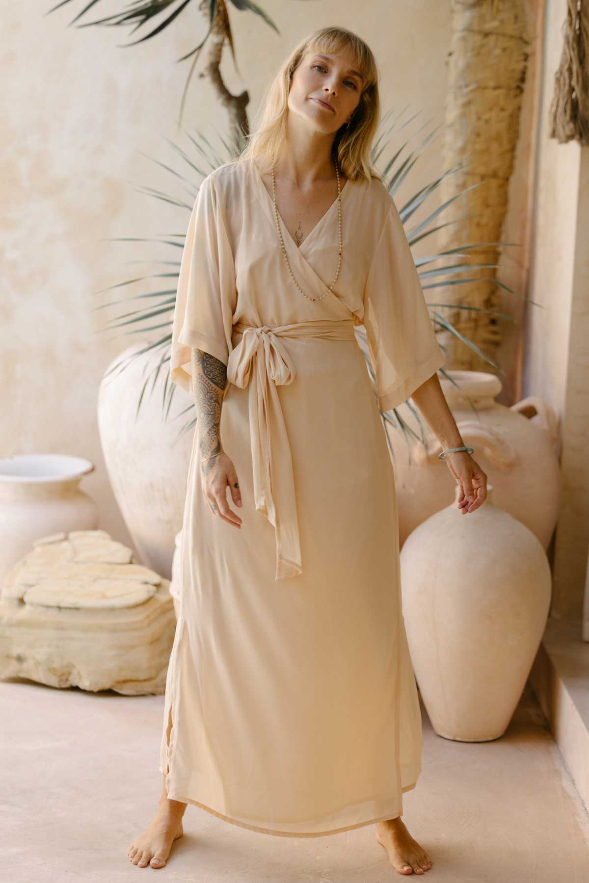 A woman with long blonde hair stands barefoot on a beige floor, wearing a light beige, flowy Magdalena Gown Sheer Chiffon by Myrah Penaloza, tied at the waist. She poses in front of large, round planters and a tall plant against a neutral-toned wall, with one arm resting by her side and the other bent at the elbow.