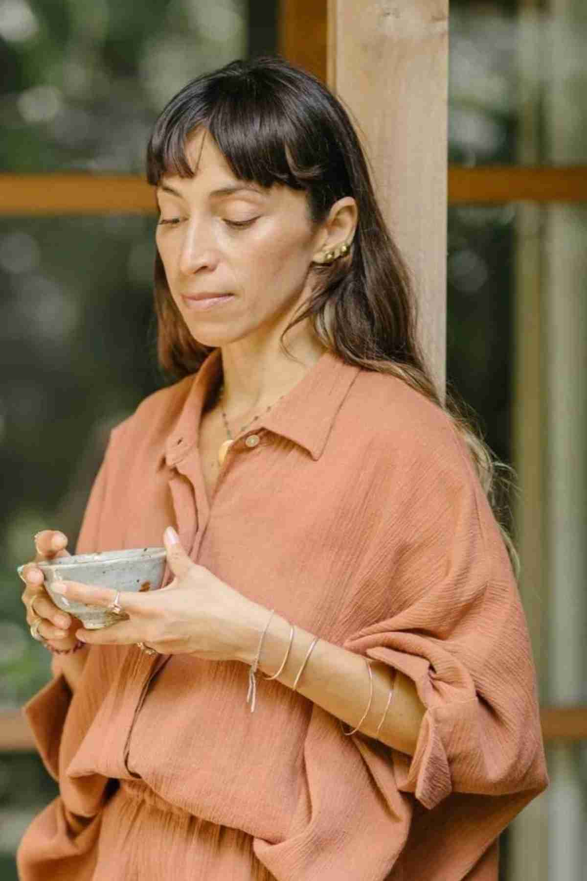 A woman with long dark hair stands outside, holding a small bowl in both hands. Dressed in the Suka Button Down Top by Myrah Penaloza, made from plant-dyed fabric in a loose camel color, she exudes a calm and reflective mood, embodying the essence of sustainable fashion.