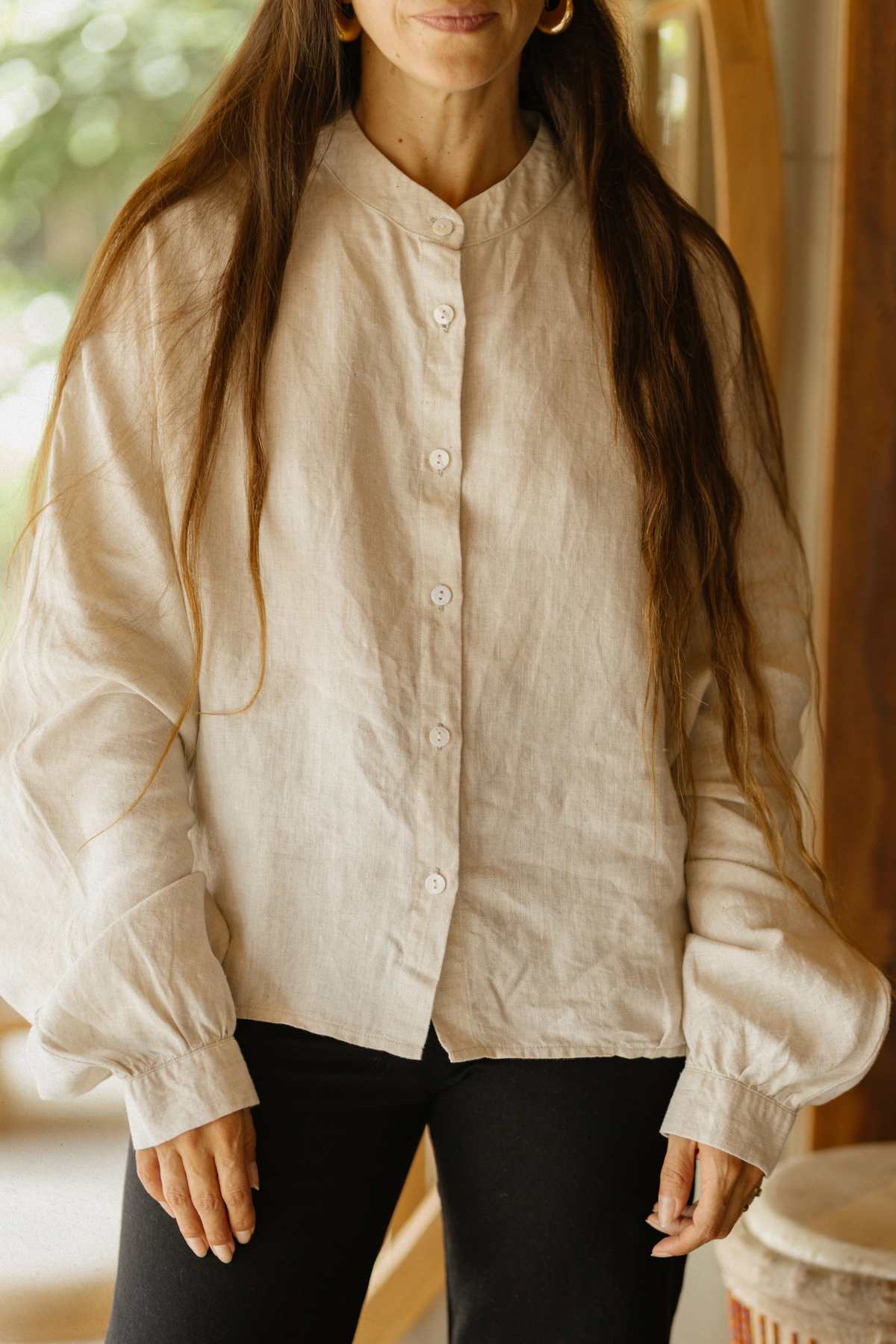 A person with long hair wearing the Cardenal Linen Top by Myrah Penaloza and black pants stands indoors. The background is softly blurred, highlighting wooden beams and a hint of greenery outside.