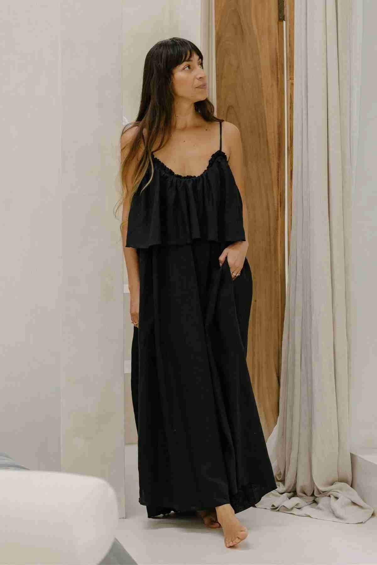 A woman stands barefoot in a relaxed pose, wearing the Linen Muse Gown by Myrah Penaloza. The loose-fitting, sleeveless black dress features adjustable straps and a low-back design. Her long, dark hair falls over her shoulders as she looks thoughtfully to the side. She is positioned near a wooden door, framed by light-colored curtains.