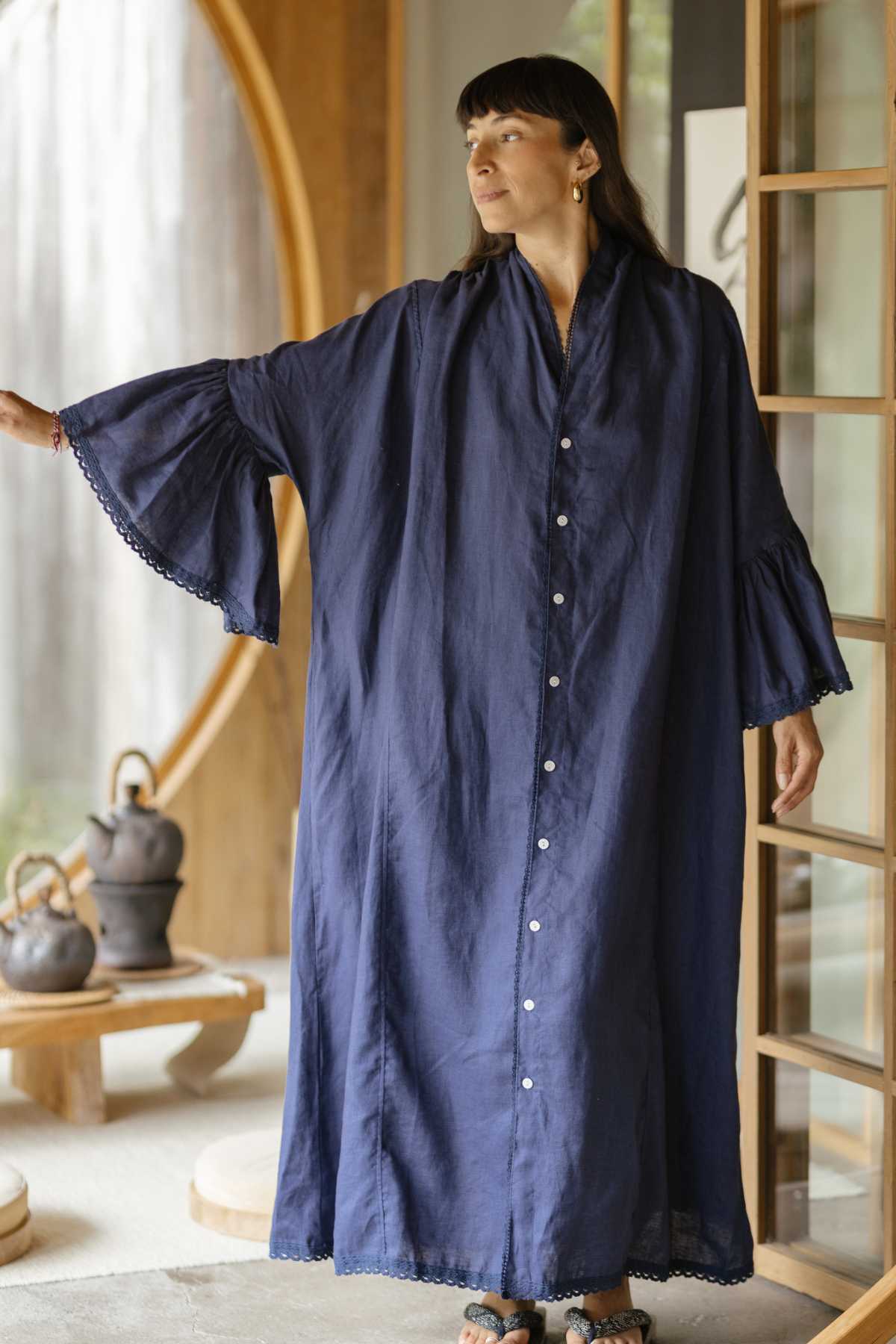 A person stands indoors dressed in the elegant The Juliette Linen Gown by Myrah Penaloza, featuring wide, flowing sleeves in a rich navy blue hue. They glance to the side, framed by a wooden circular door with a table adorned with teapots in the background.