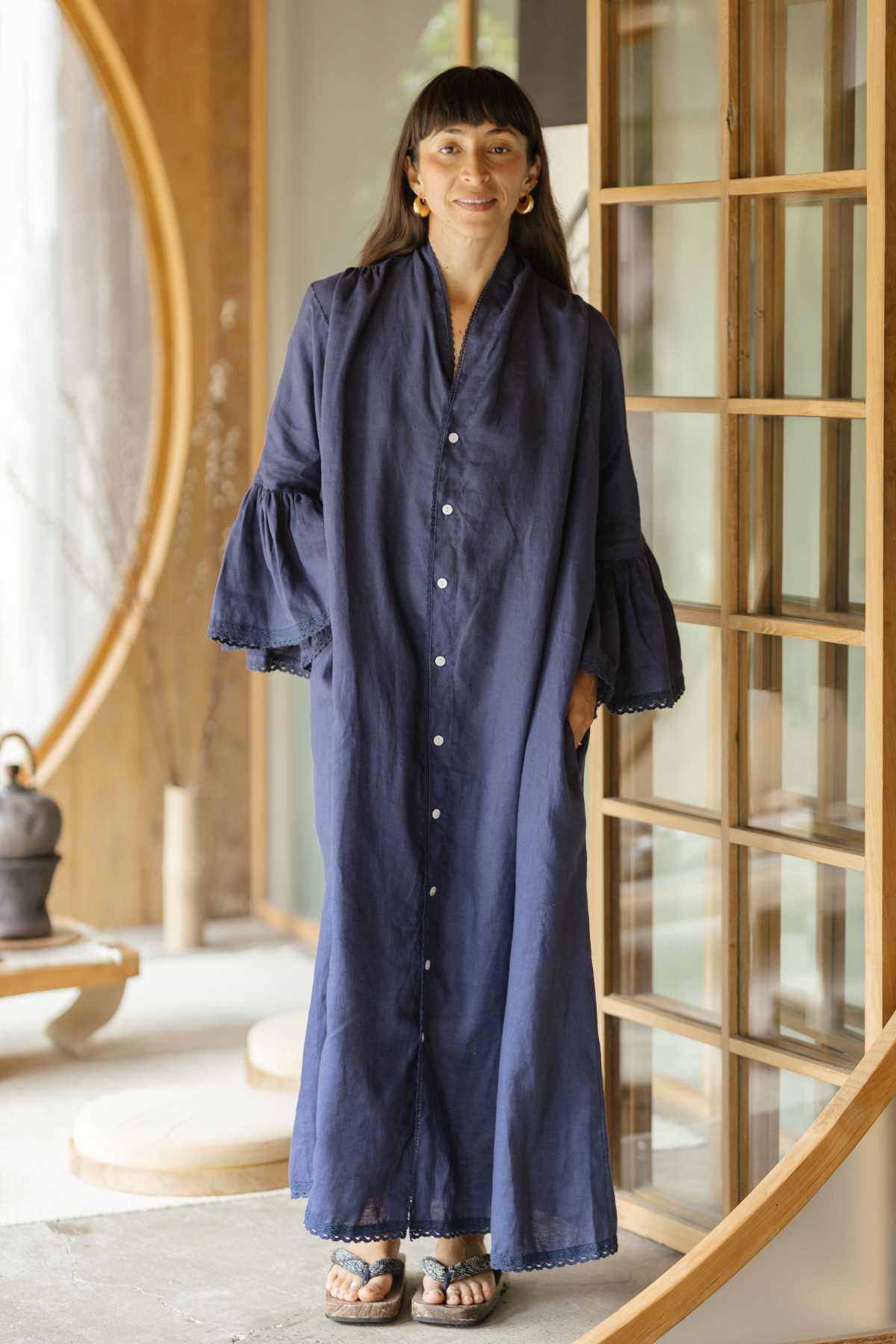 A person in a Kyoto Duster, made to order by Myrah Penaloza, stands indoors near wooden sliding doors. The long, navy blue dress features button detailing and bell sleeves with delicate cotton lace accents. They are wearing sandals and have shoulder-length hair, smiling softly at the camera. In the background, a teapot is visible on a table.