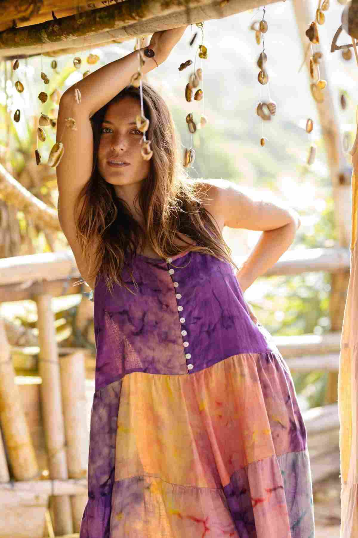 Wearing the La Mexicana Playsuit by Myrah Penaloza, a woman in a vibrant, tie-dye outfit with adjustable straps leans against a wooden railing decorated with hanging shells. Her long hair cascades down as she gazes directly at the camera, surrounded by wooden structures and lush greenery that capture the essence of a breezy summer day.