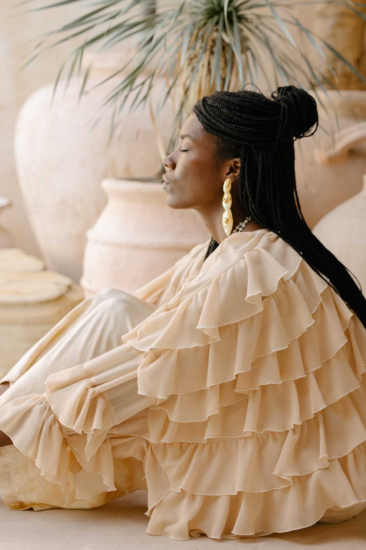 A woman wearing the Anusara Bloom - Made To Order top by Myrah Penaloza and long earrings sits peacefully with her eyes closed. Her long, braided hair complements the Nature-Inspired Couture setting as she is surrounded by large ceramic pots and a plant, creating a serene and natural backdrop.