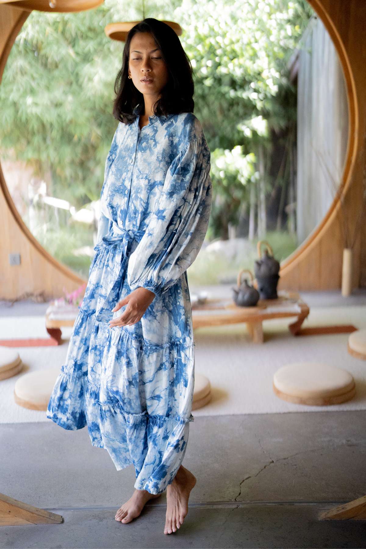 Standing barefoot indoors, a person is wearing the La Dama Long Sleeves Ruffles gown in hand tie-dyed indigo by Myrah Penaloza. Positioned near a circular window with views of lush greenery, the room—featuring wooden seating and decor—exudes a serene, renaissance-inspired elegance.