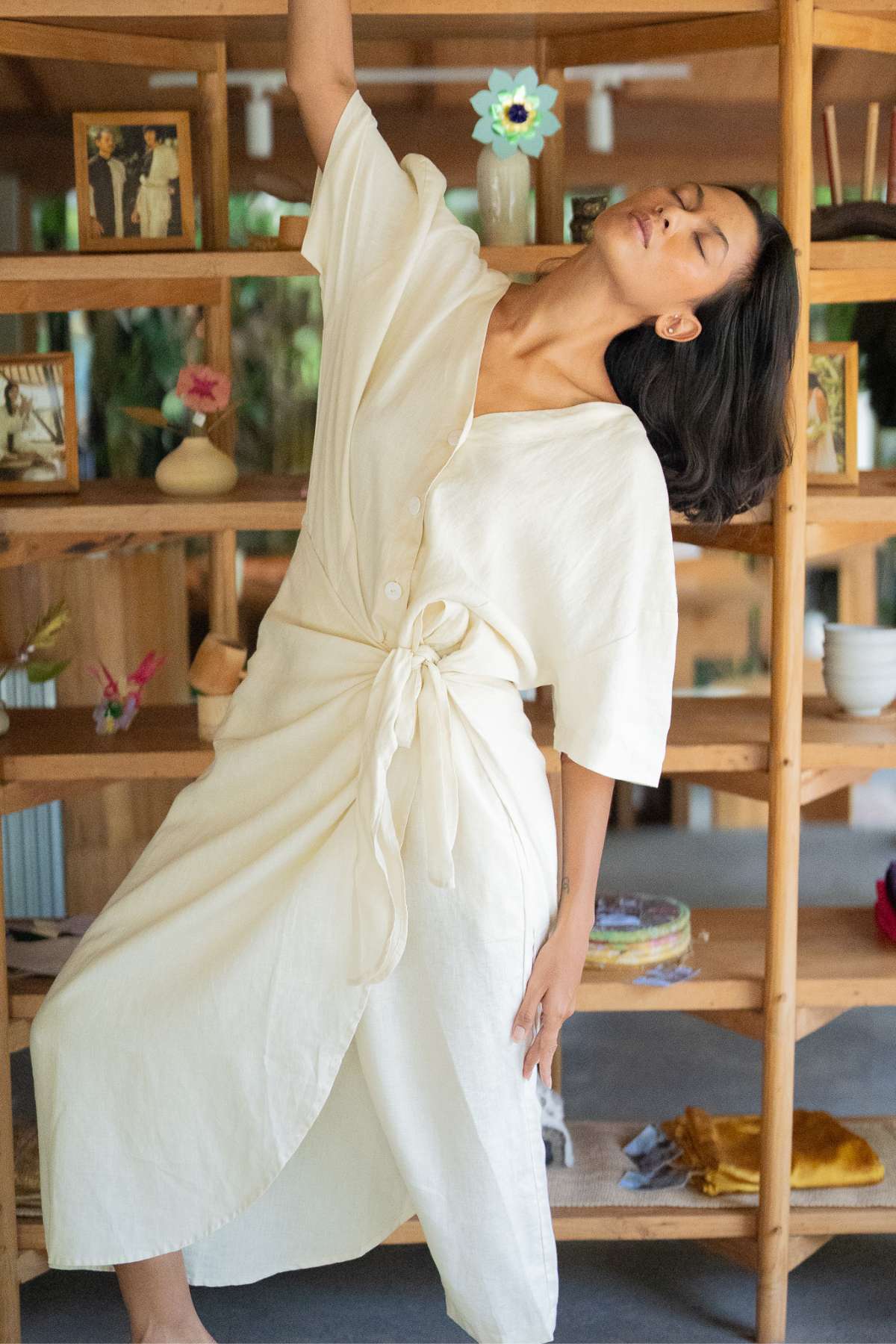 A woman wearing the Temple Wrap Playsuit Linen by Myrah Penaloza, reminiscent of a sarong, poses gracefully in front of a wooden shelving unit adorned with small plants, flowers, and framed photos. The soft lighting creates a warm, serene atmosphere that echoes the tranquility of Balinese culture.