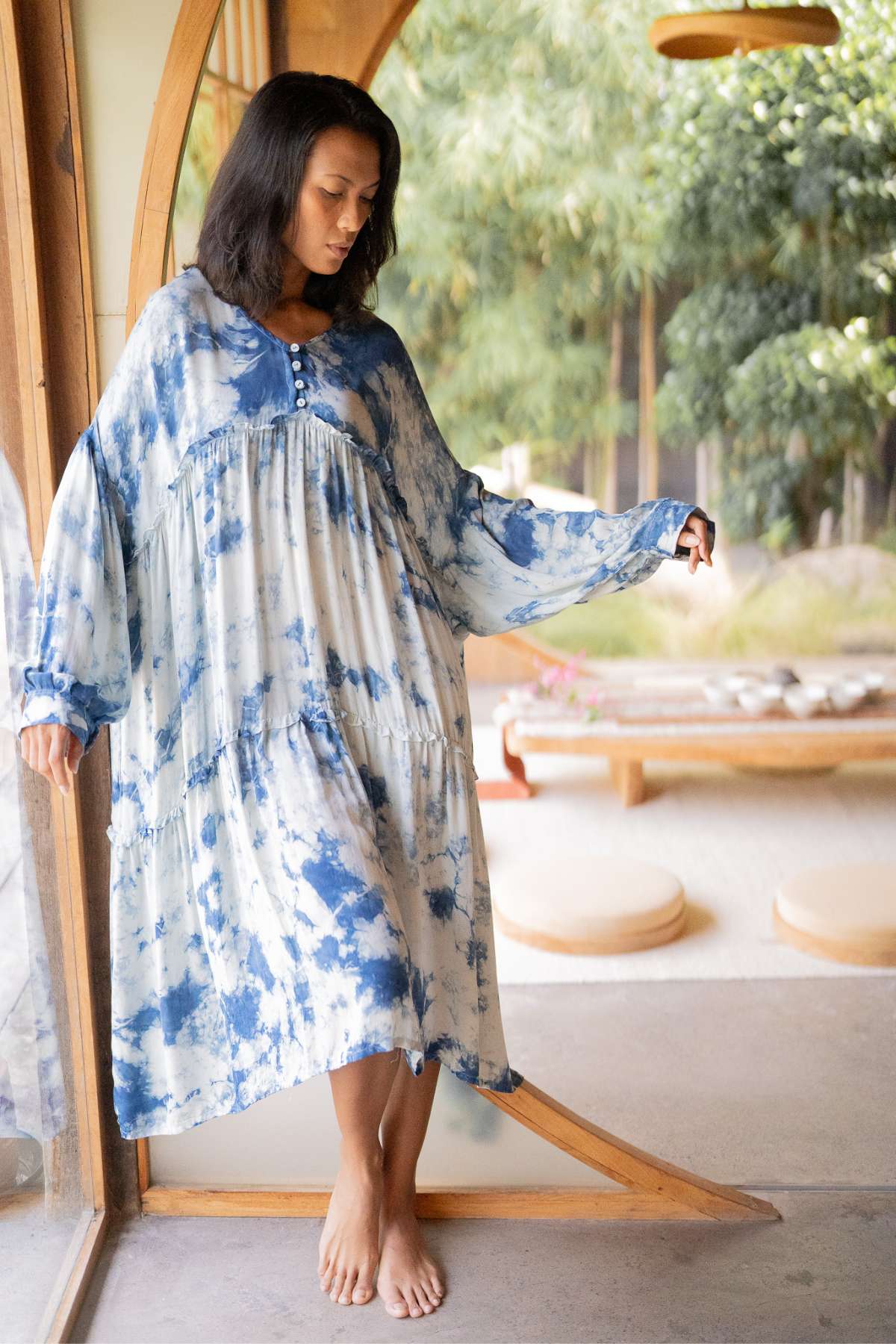 A woman stands by a large window, gazing down in the flowing Indigo Eva Gown made from 100% silk by Myrah Penaloza. The space is bathed in natural light, showcasing a wooden floor and lush plants outside. Her gown, with its oversized fit, billows gently around her, adding elegance to the serene atmosphere.