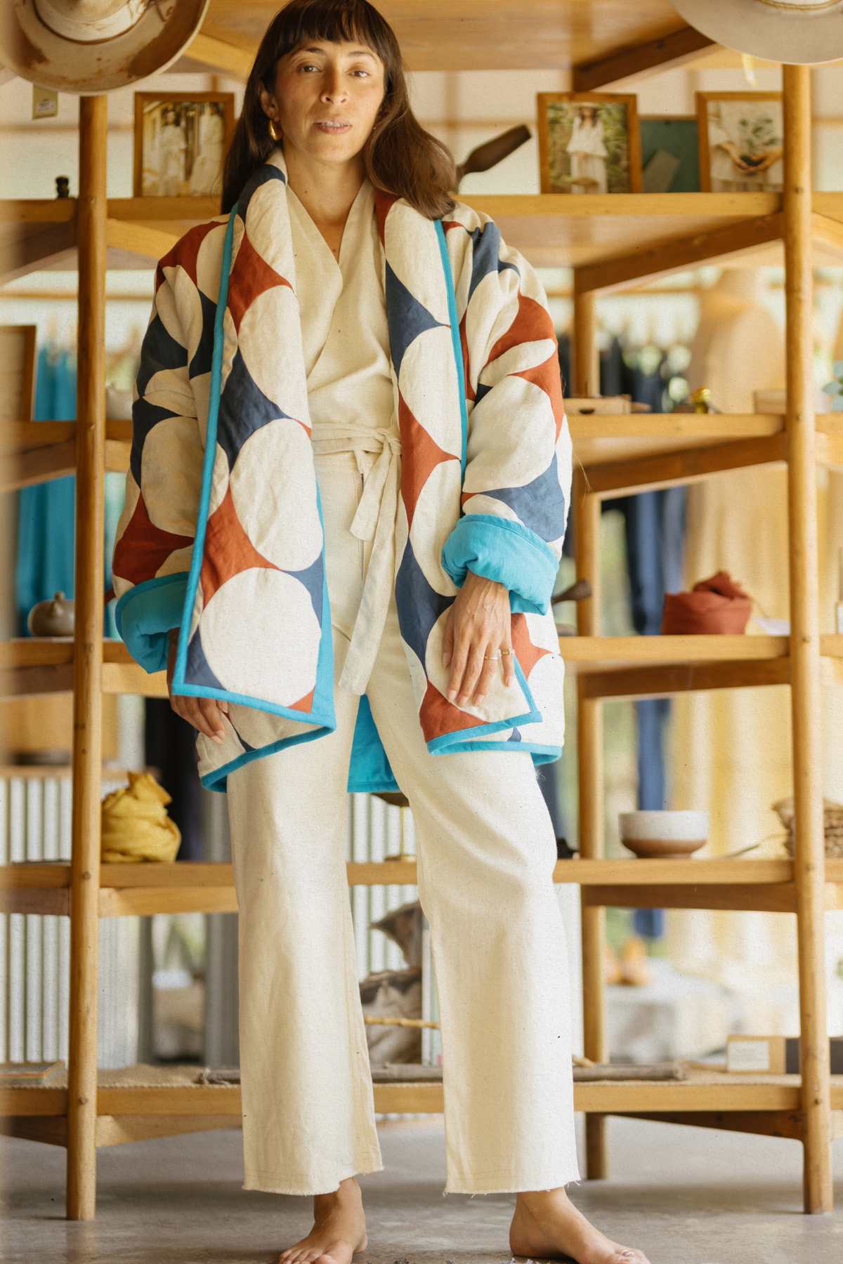 A person stands barefoot in a room, clad in a white outfit complemented by the Javi Quilted Jacket from Myrah Penaloza. The jacket boasts a vibrant geometric pattern of large red, blue, and white circles. They are surrounded by wooden shelves displaying various textiles and decor items, epitomizing the charm of unisex clothing.