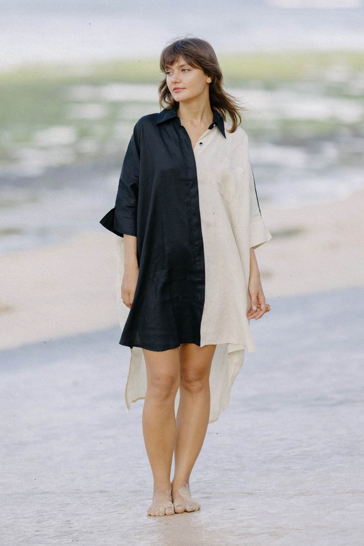 A woman stands barefoot on a beach, wearing the Linen Suka Button Down Long Top by Myrah Penaloza. This loose-fitting dress, made from sustainable fabric in a half-black-and-half-white design, features buttons down the front. She gazes to the side with soft waves in her hair and a calm expression. The background reveals a sandy shore and a glimpse of the ocean.