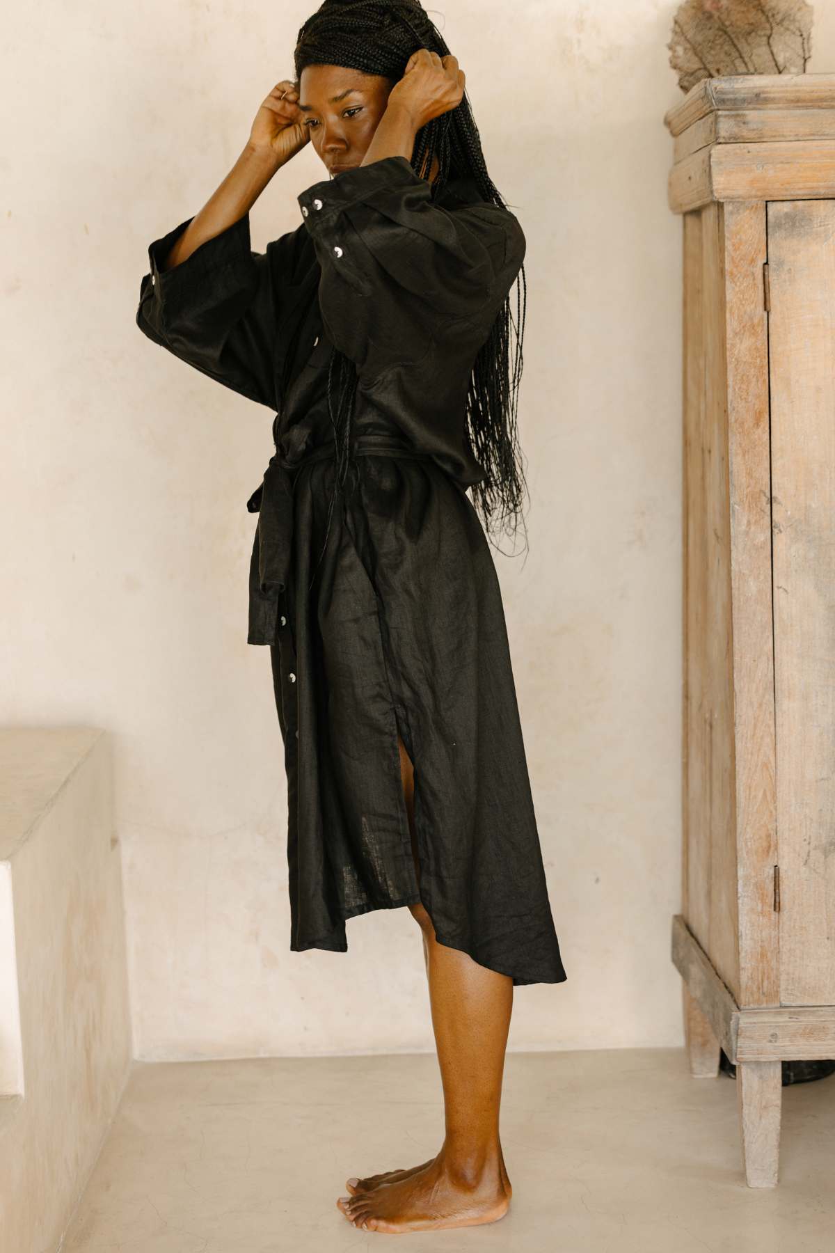 A person stands barefoot on a light-colored floor, adjusting their long black braids. They are wearing the Nidra Full Length Linen Duster, a sustainable clothing piece by Myrah Penaloza, which is a black, long-sleeved dress with a belted waist. A wooden cabinet is partially visible in the background.