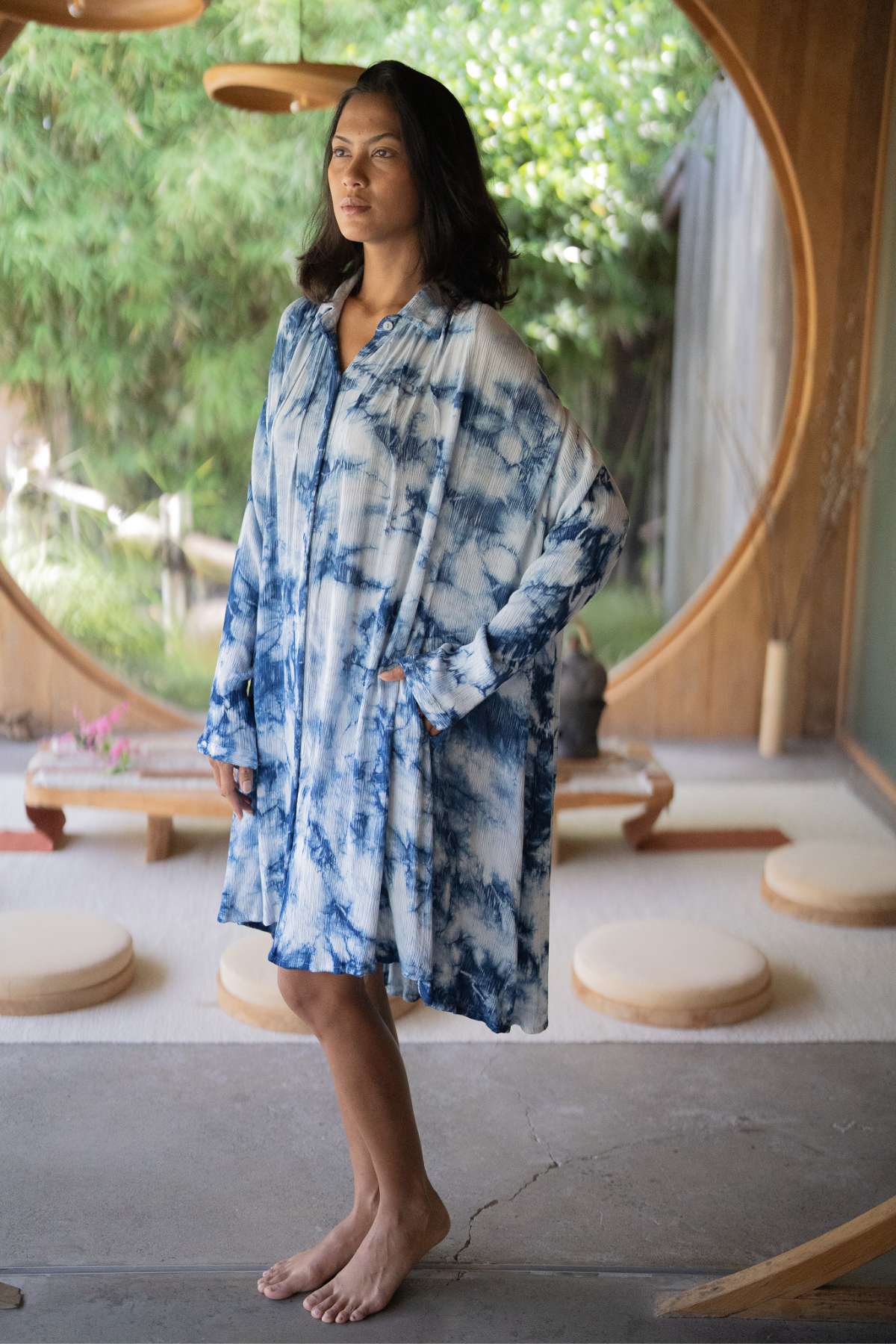 A person stands barefoot indoors, dressed in the Indigo Dreams Kundalini Gown Short by Myrah Penaloza. The gown features a flowing design made from bamboo rayon in a blue and white tie-dye pattern. In the background, circular windows offer a view of lush greenery, enhancing the peaceful, natural ambiance.