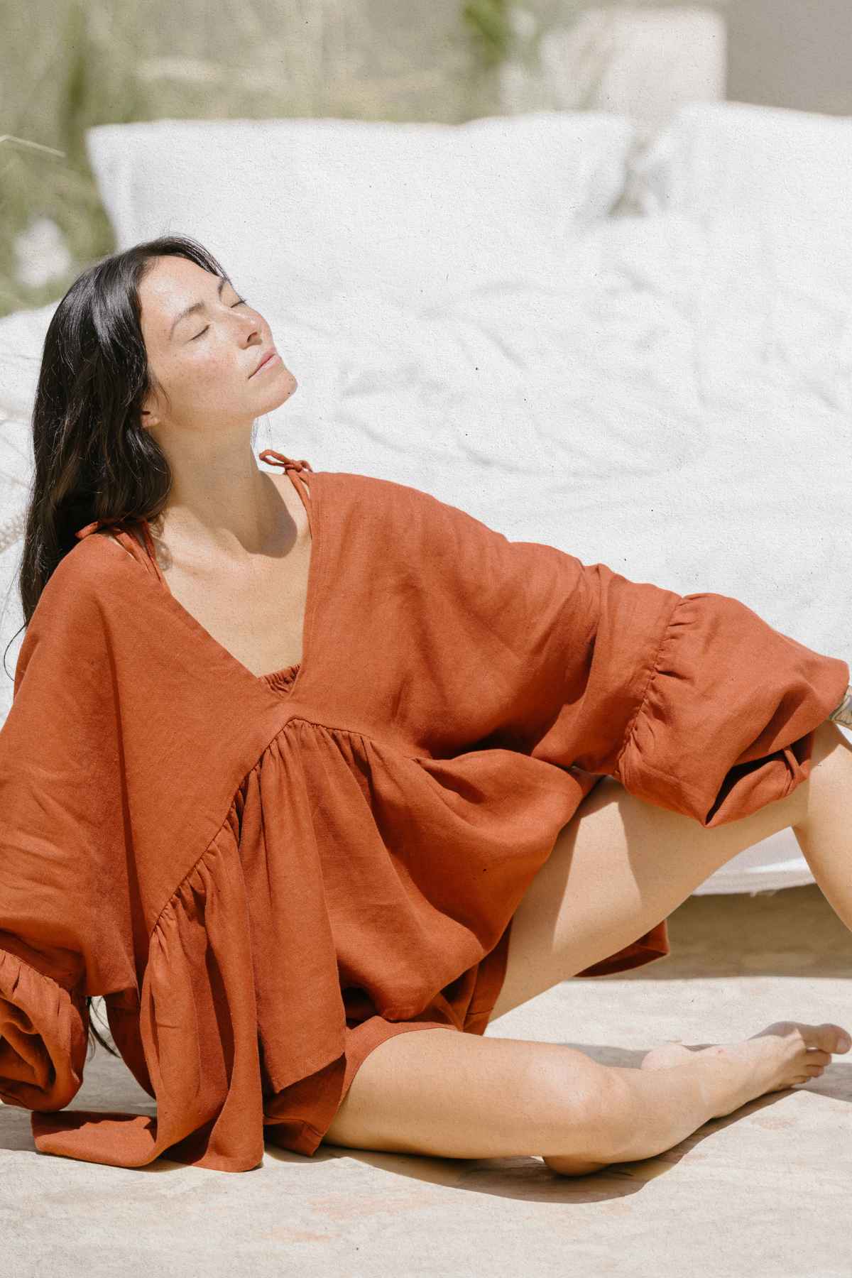 A woman sits outdoors on a sunny day, eyes closed and head tilted upward, possibly enjoying the warmth of the sun. She wears a loose, rust-colored Eva Linen Top by Myrah Penaloza with billowy sleeves that look like flowering sleeves, and her long, dark hair cascades over her shoulders. White fabric is visible in the background.