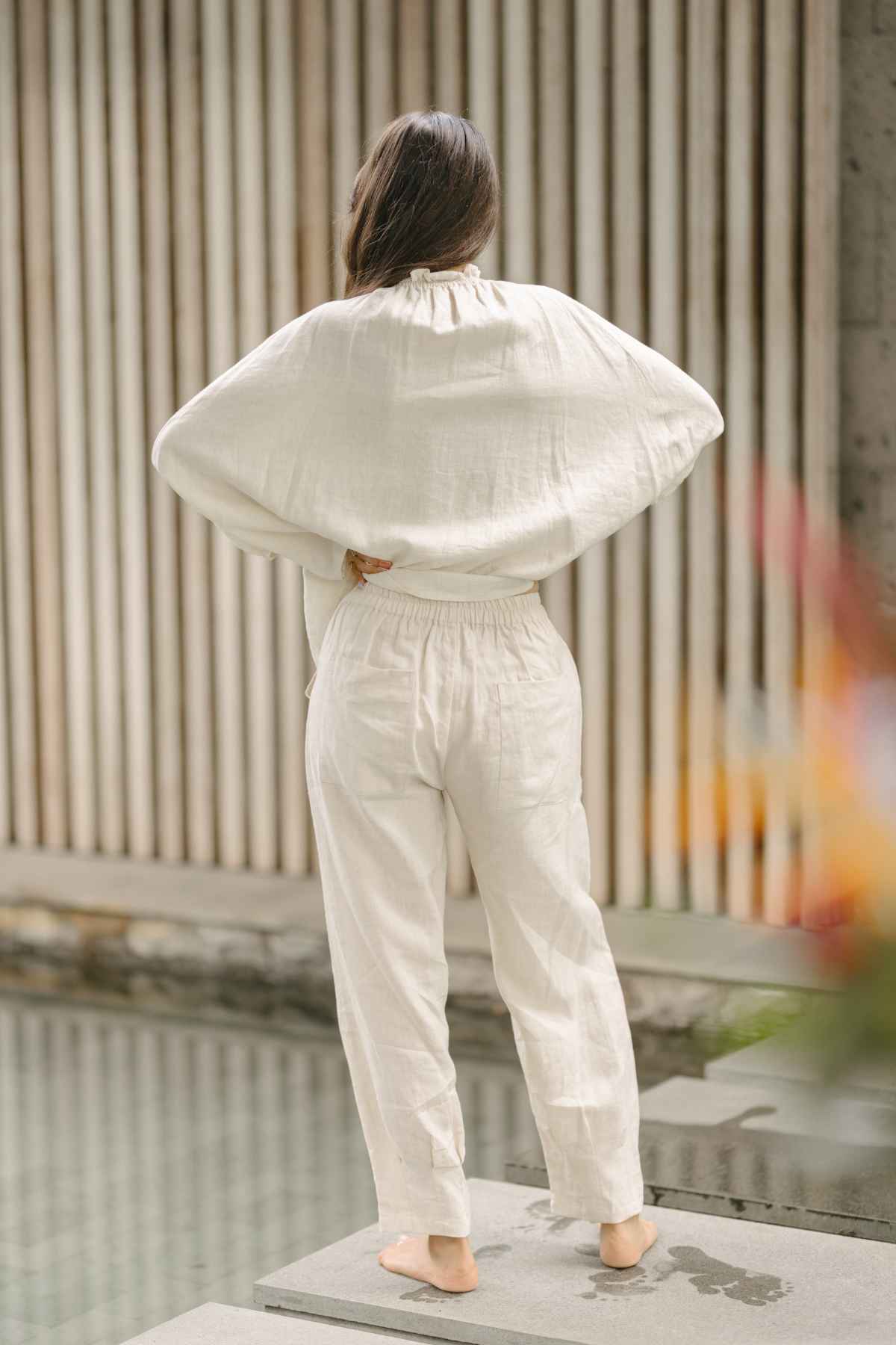 A person stands barefoot on a flat surface, facing away from the camera. They are wearing a loose-fitting, long-sleeve white top and matching Myrah Penaloza's Linen Chajin Pant. The background includes vertical wooden slats and a water feature, creating a serene scene perfect for any versatile wardrobe.