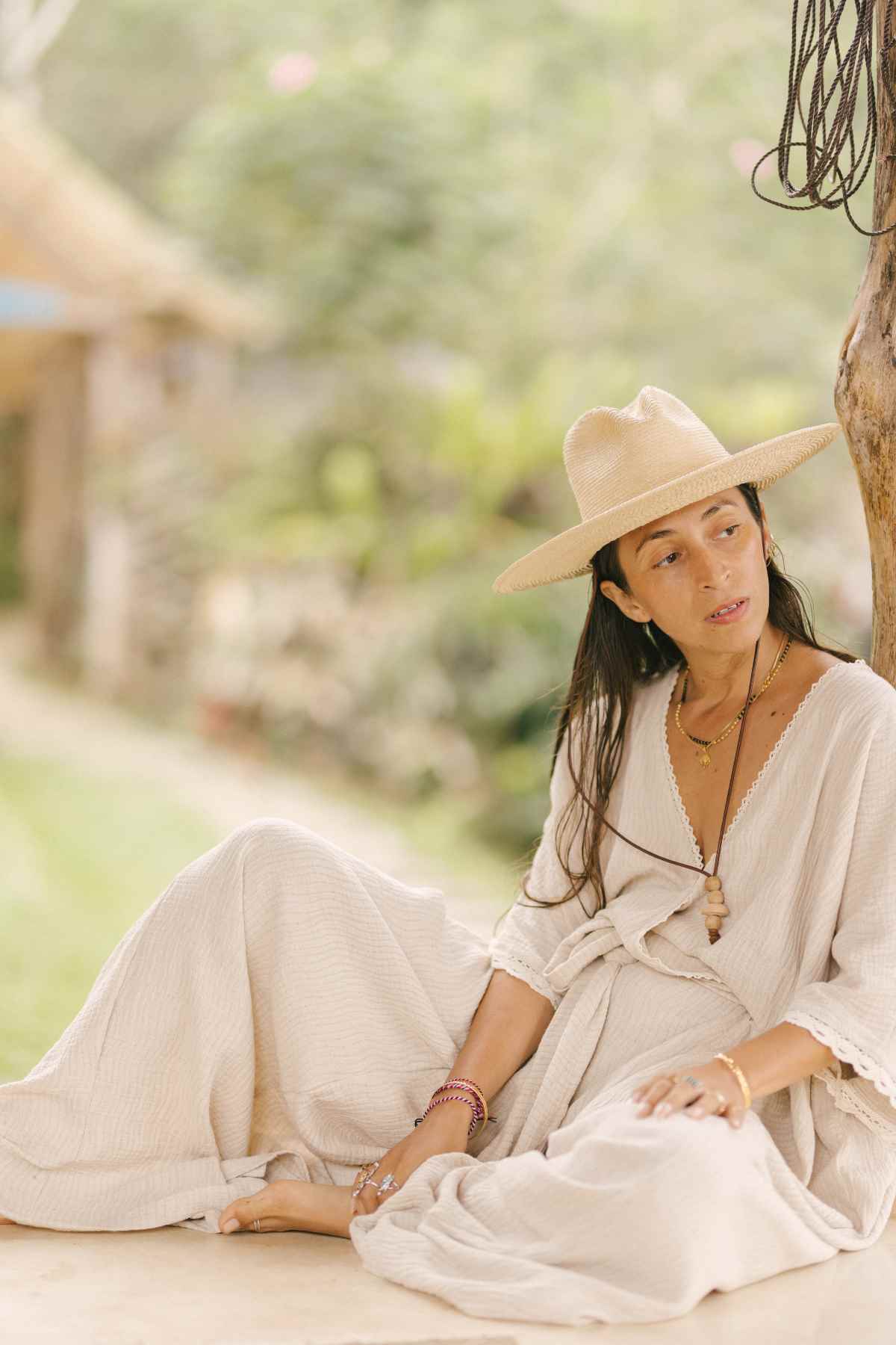 A person wearing a wide-brimmed straw hat and a light, loose-fitting Limited Edition Hand Embroidered River Playsuit by Myrah Penaloza sits on the ground outdoors. They look to the side while holding their hat. The background is a lush, green area with what appears to be a thatched-roof structure.