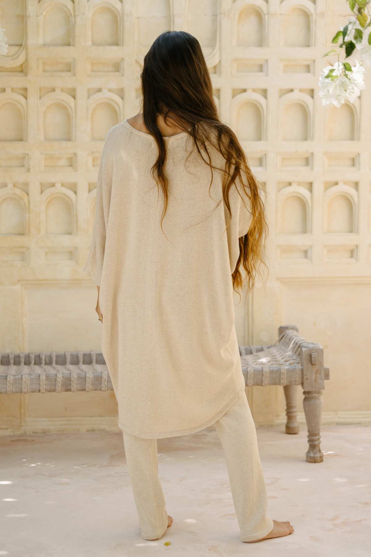 A person with long, wavy hair stands with their back to the camera, wearing a light beige, flowing Butterfly Tunic Cotton Knit from Myrah Penaloza and matching pants. They are outdoors near a decorative, arched wall, with a rustic bench to their right and greenery hanging overhead.