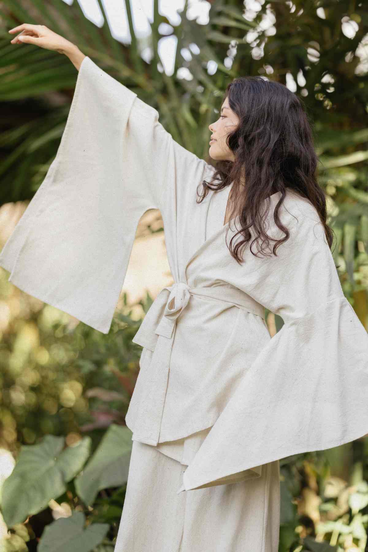 A person with long, wavy hair is standing outdoors among lush green foliage. They are wearing the light-colored Dewi Set (3 of a kind) - Pre-order by Myrah Penaloza, a belted kimono made of hypoallergenic linen cotton crepe with wide sleeves, and have one arm raised, looking to the side. The atmosphere is serene and natural.