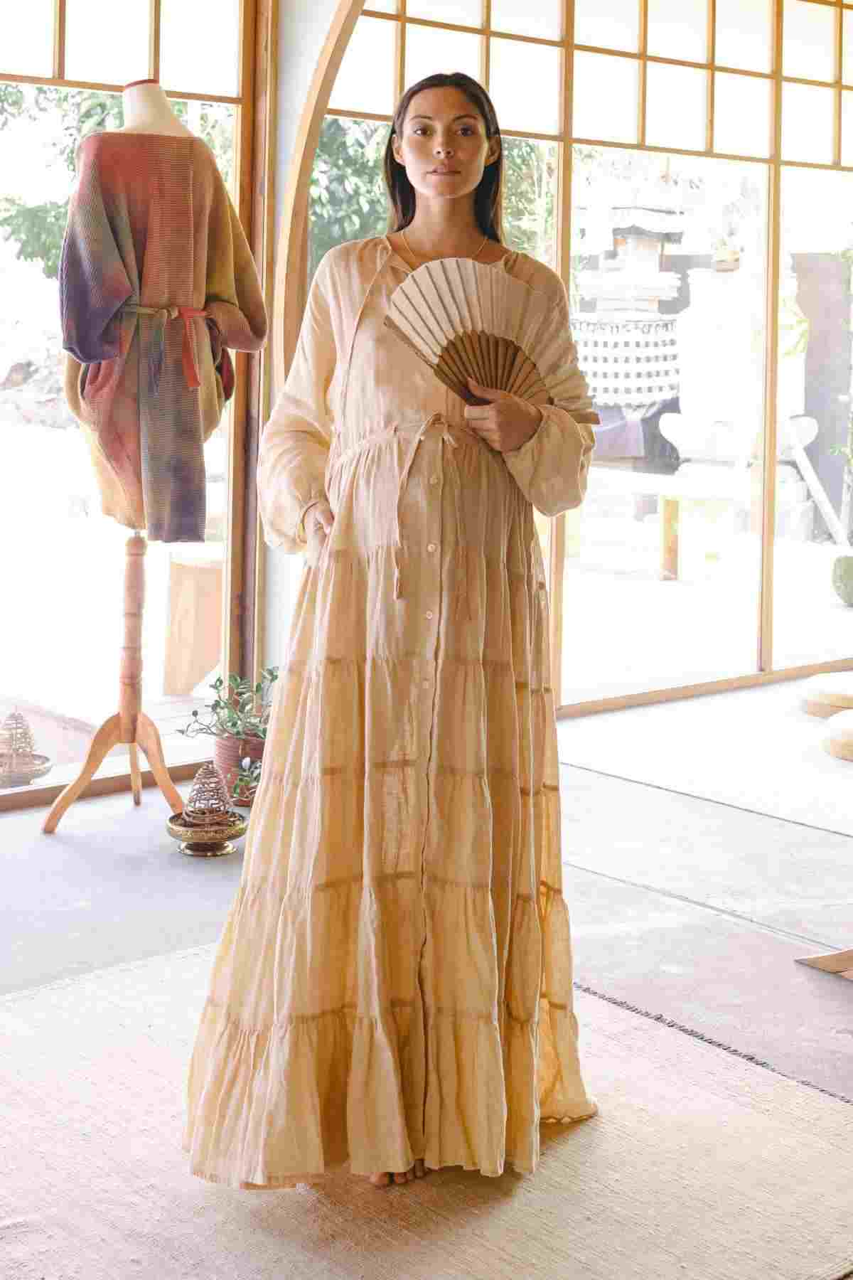 A woman stands indoors wearing the La Mexicana Gown by Myrah Penaloza, showcasing its long, flowing beige design with an adjustable waist, while holding a fan. Behind her is a mannequin dressed in a colorful ensemble. The room, featuring large windows, radiates a serene and sunlit ambiance.