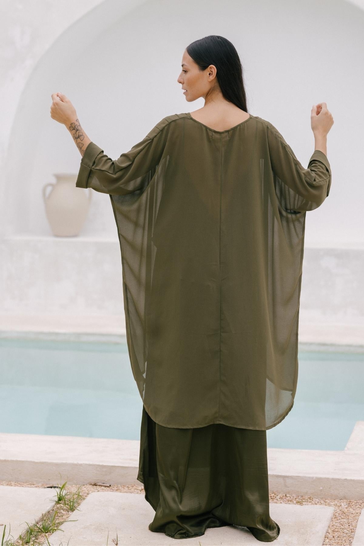 A woman in Myrah Penaloza's luxurious cape, crafted from 100% silk in olive green, stands near a pool. Her long dark hair cascades down her back as she raises her arms slightly toward a white arch in the background. The elegant fabric whispers grace as it drapes beautifully around her.