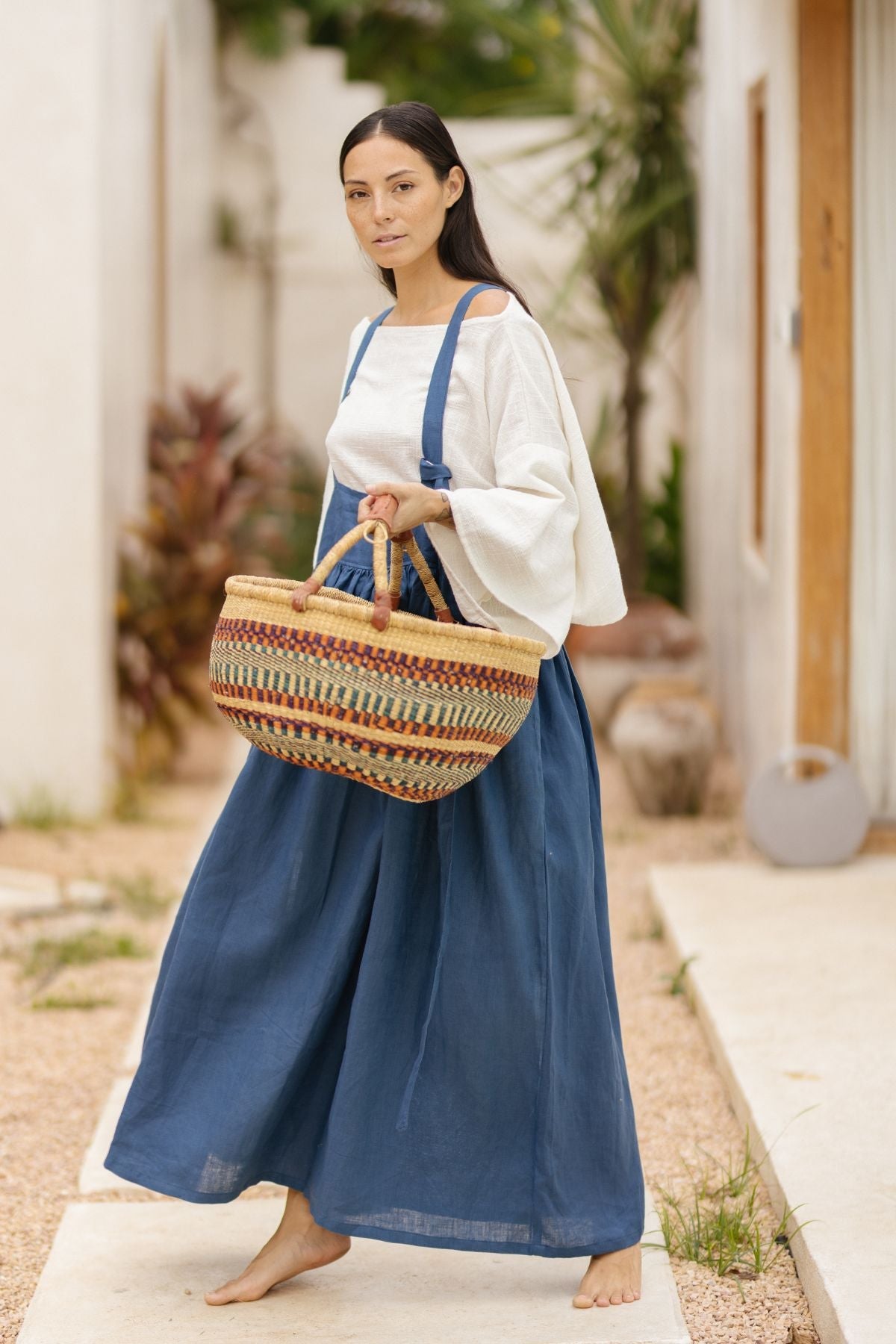 A barefoot woman with long dark hair stands outdoors on a light surface. She wears a white blouse and the May May Overall Skirt (Made To Order) by Myrah Penaloza. She carries a large, colorful woven basket with both hands. There are plants and buildings in the background.
