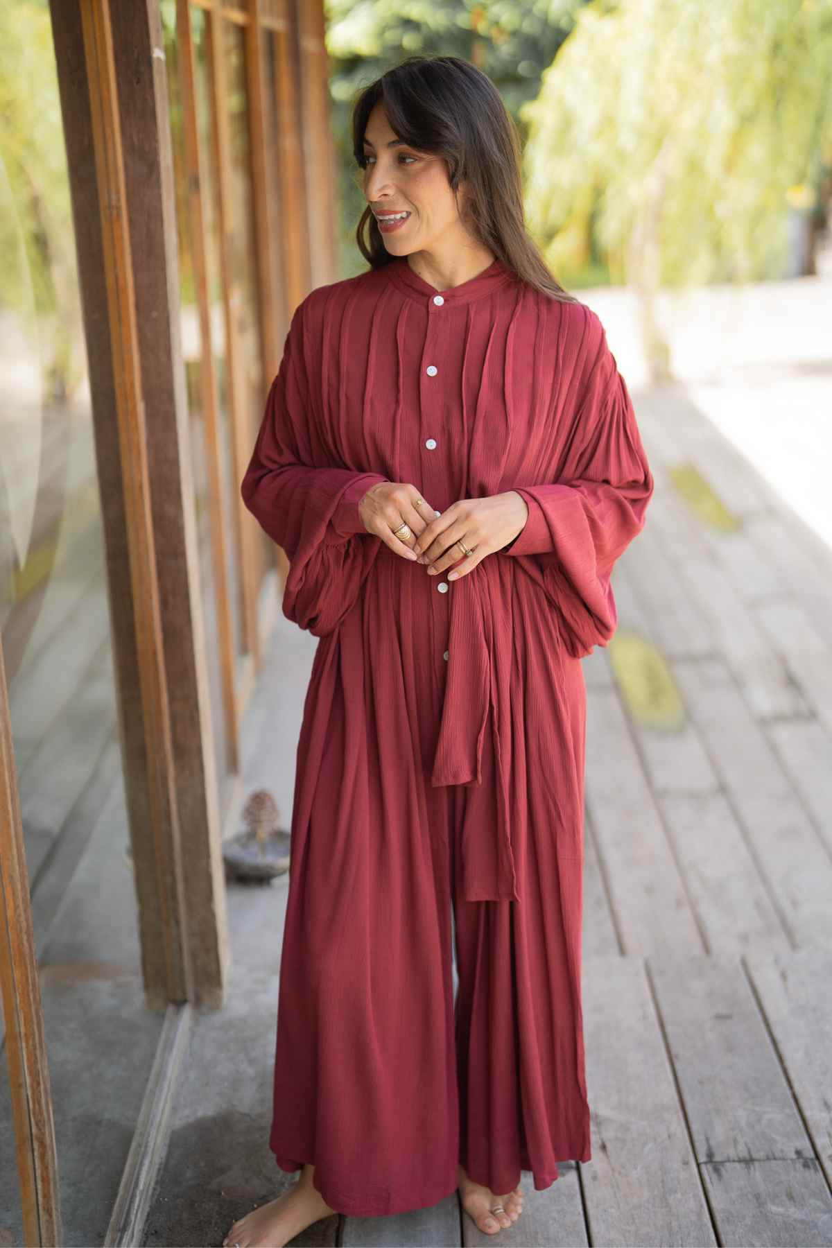 A woman wearing a long-sleeved, flowing Kundalini Gown Playsuit Edition from Myrah Penaloza stands barefoot on a wooden deck. She is smiling and looking to her right, with her hands clasped in front of her. Behind her, there is a large glass window through which greenery can be seen outside.