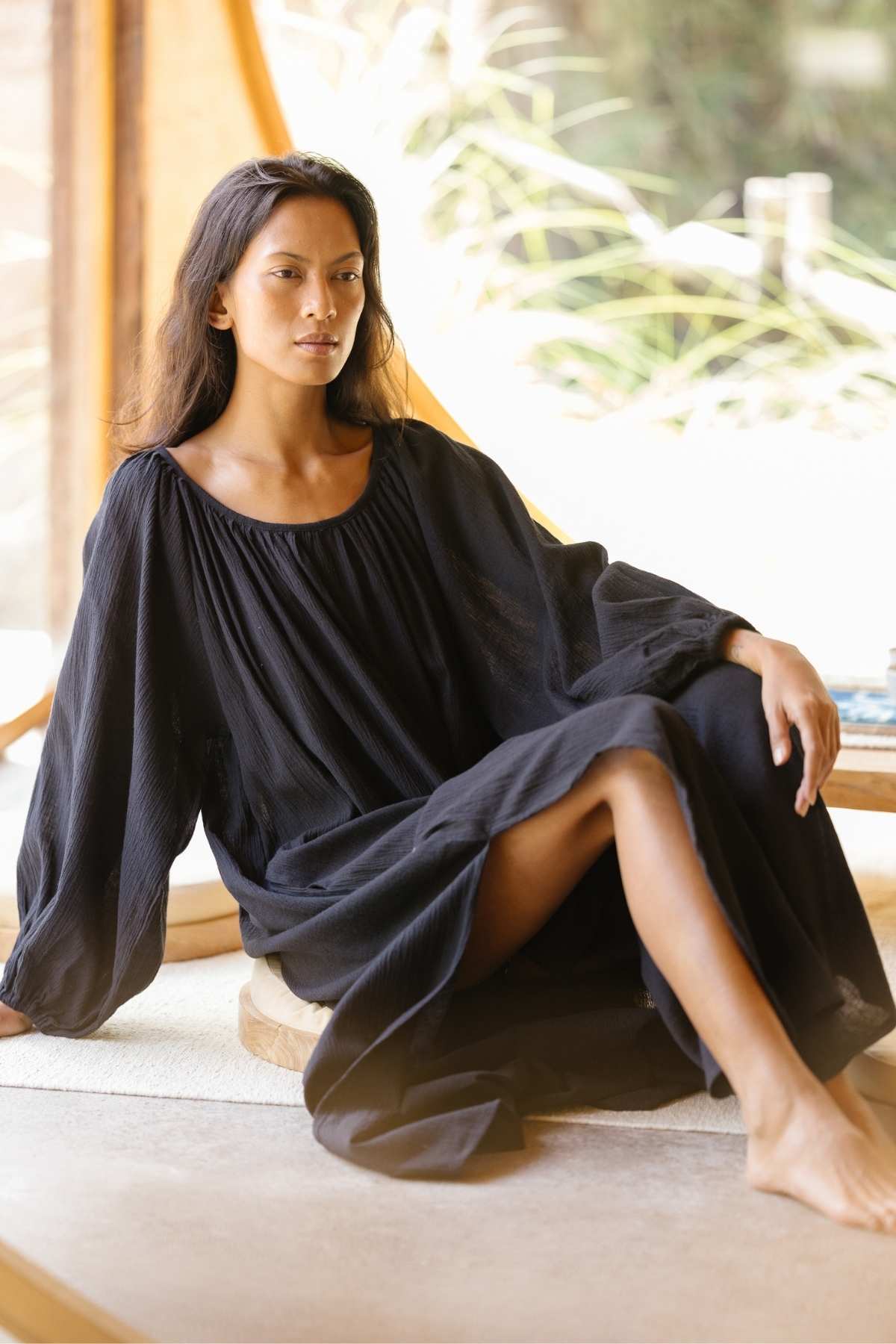 A woman with long dark hair sits comfortably on a cushion in a naturally lit room. She is wearing a loose, flowing Alchemy Gown (Crinkle Linen Cotton) by Myrah Penaloza with long sleeves. Sunlight filters through large windows behind her, casting a soft, warm glow into the space, enhancing her aura of a modern mystic.