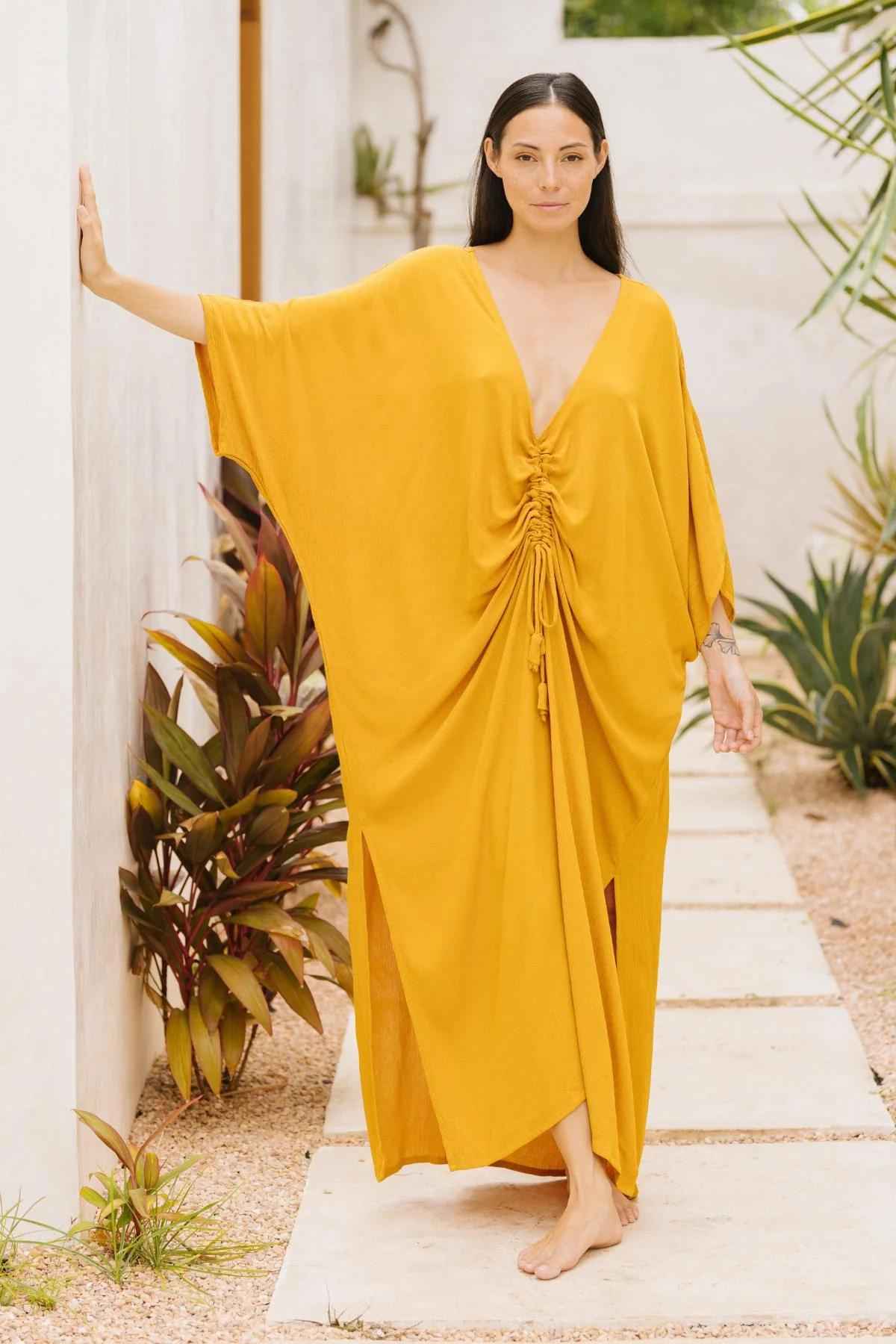 A woman stands barefoot on a stone pathway beside a white wall, embodying sophisticated fashion in a Myrah Penaloza Limited Edition Virgo Kaftan made of luxe bamboo rayon. With long dark hair and one hand resting on the wall, she showcases the kaftan's deep V-neckline and ruched front. Plants are visible in the background.