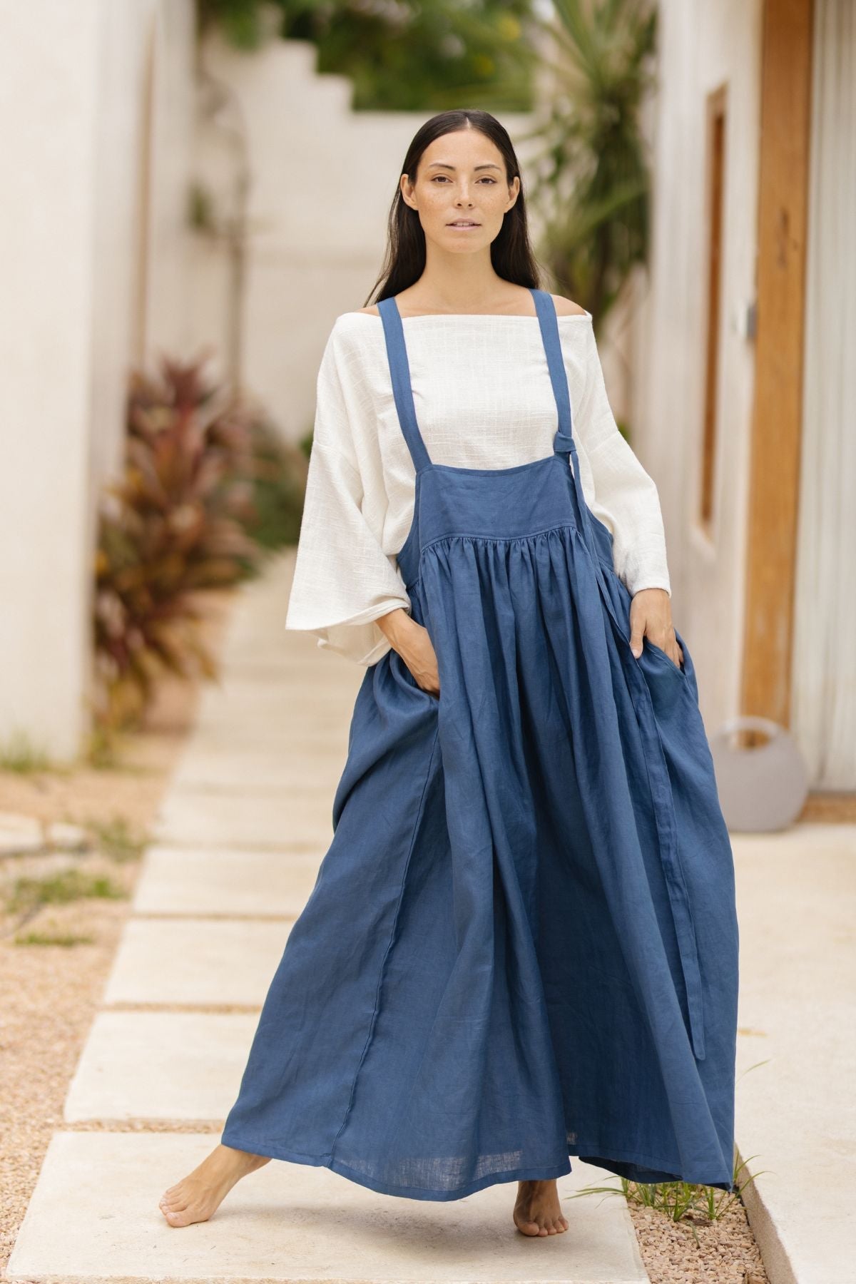 A woman stands outdoors on a stone walkway lined with greenery, wearing the May May Overall Skirt (Made To Order) by Myrah Penaloza. The skirt is a long blue overall design made of 100% linen, paired with a white blouse and her hands in the pockets. The background features a wall adorned with plants and an architectural structure.