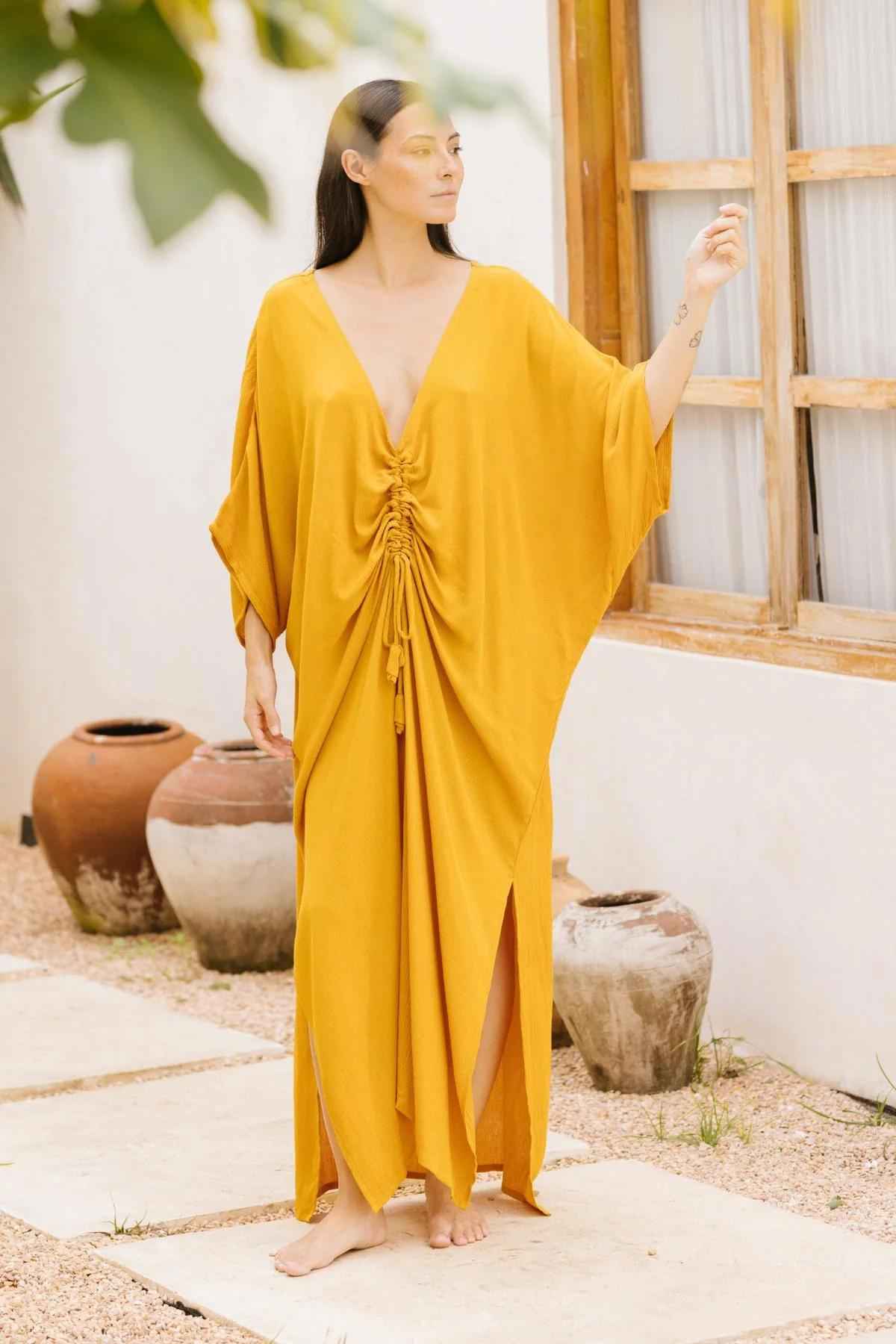 A barefoot woman stands on a stone path outside a white building, adorned in Myrah Penaloza's Limited Edition Virgo Kaftan in yellow, featuring a deep V-neck and gathering at the front. Holding a leaf with one hand, she gazes thoughtfully to the side while clay pots adorn the background, enhancing the sophisticated fashion scene.