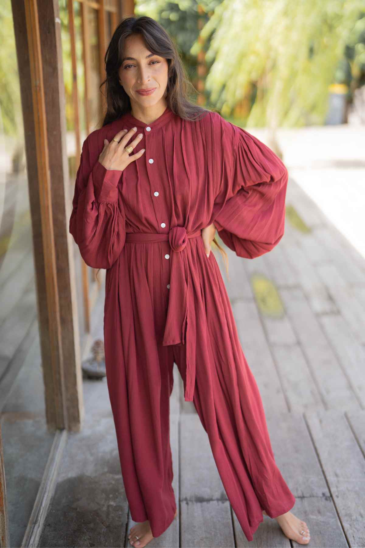 A person stands outside in a relaxed pose, adorned in the elegant Kundalini Gown Playsuit Edition from Myrah Penaloza. This flowing, long-sleeved, belted maroon number is perfect for date night. They have long, dark hair and are smiling slightly with one hand resting gently on their chest. The background showcases a wooden structure and lush greenery.