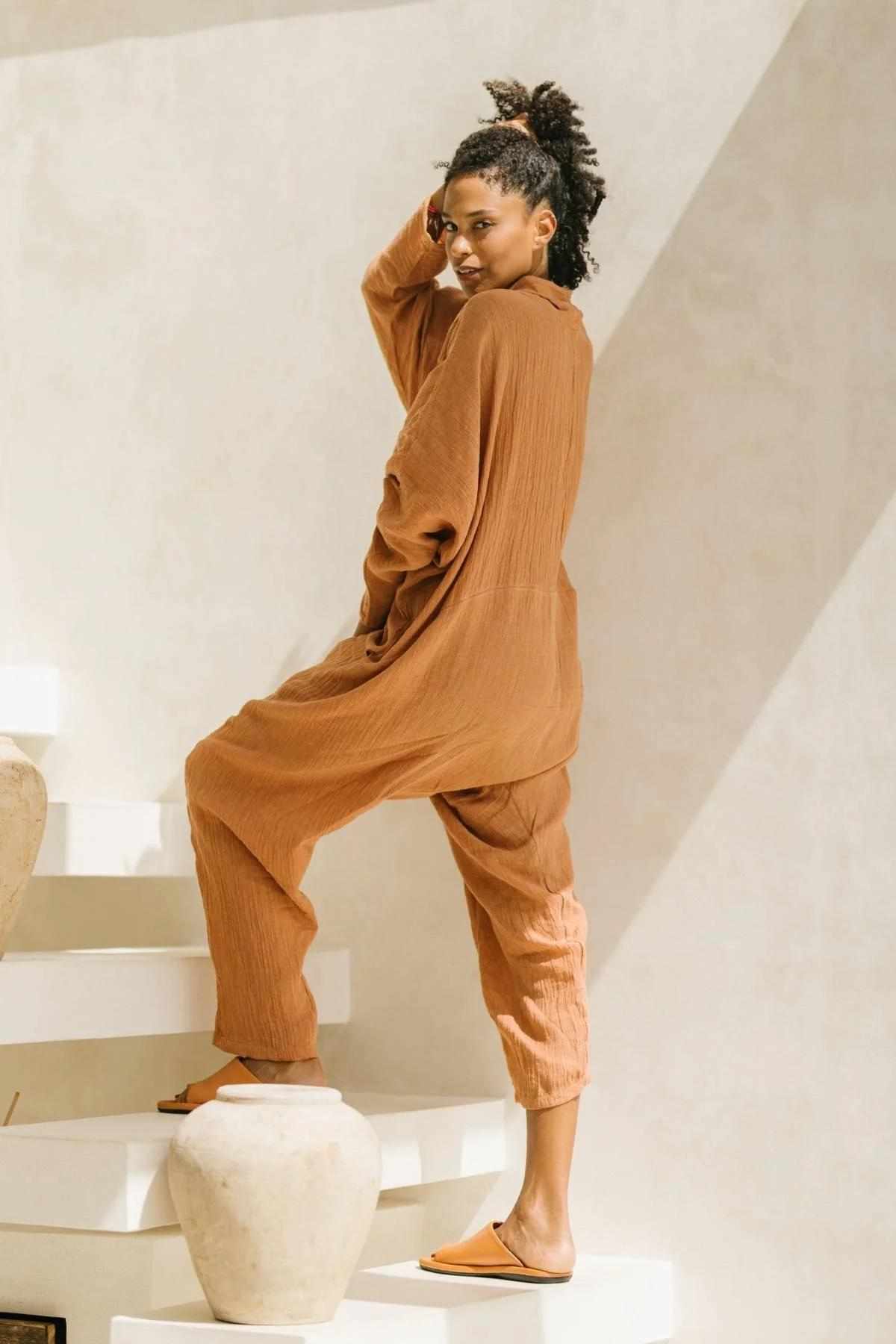 A person with an afro hairstyle poses on a white stairway in a bright setting. They're wearing the oversized Crinkle Linen Amma Playsuit by Myrah Penaloza and brown slip-on shoes, with one hand resting on their head and a confident expression. A beige vase is in the foreground.