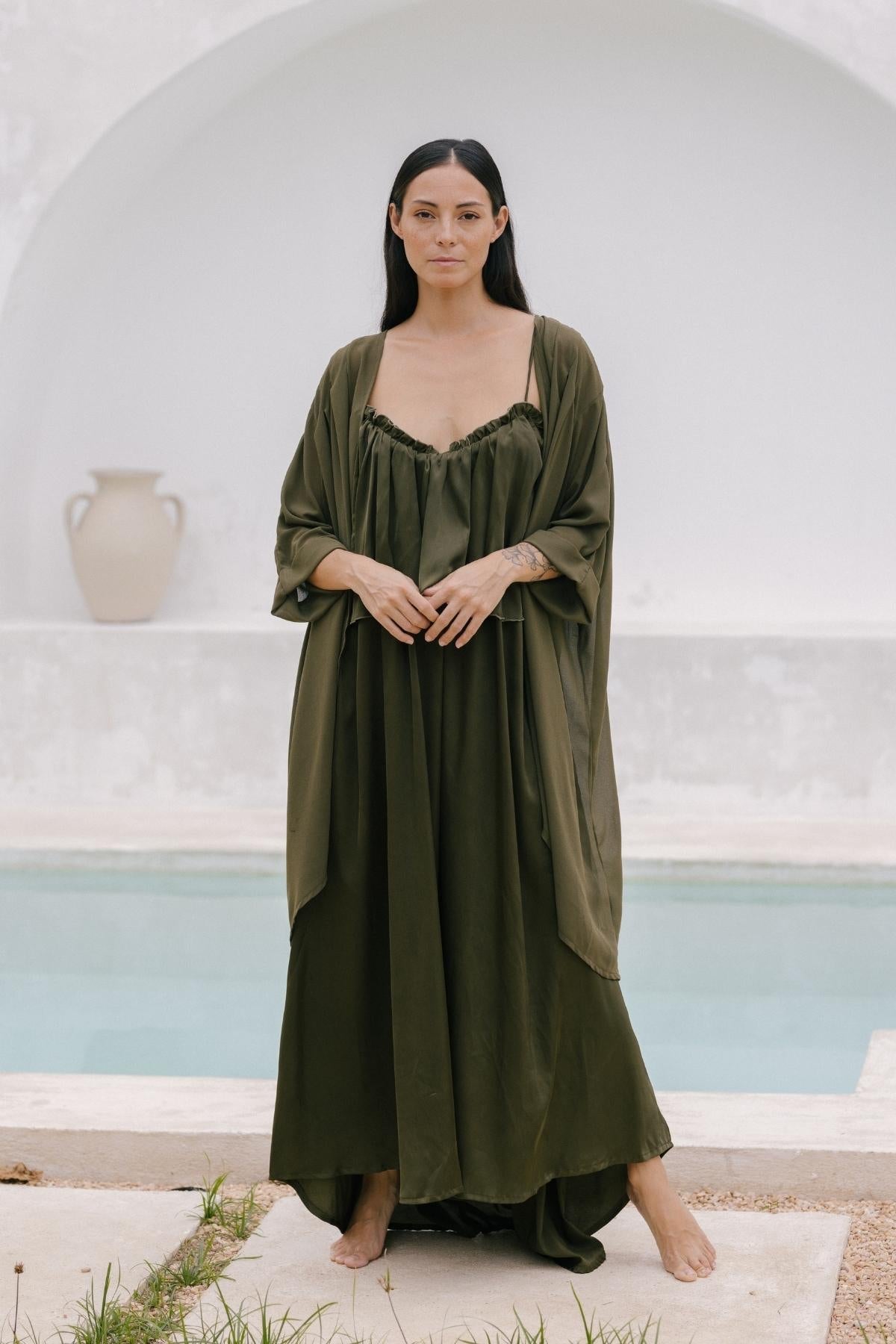 A woman stands barefoot by a pool, draped in an elegant, flowing olive green dress. Her long dark hair and serene expression are enhanced by the luxurious allure of a Myrah Penaloza silk cape against a neutral background featuring an arch and a large ceramic vase.