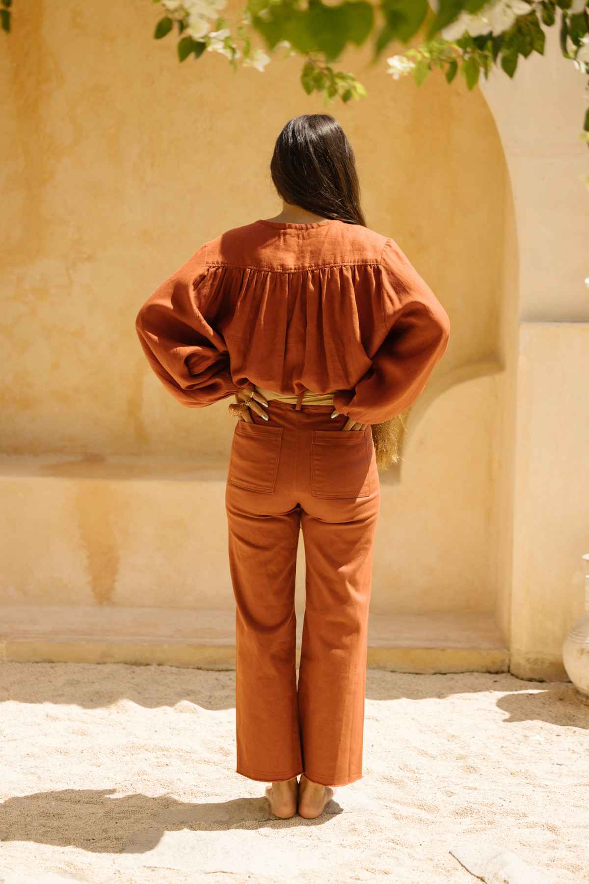 A person stands with their back to the camera, hands on hips, dressed in a loose rust-colored blouse and wearing high-waisted Vida jeans from Myrah Penaloza. The jeans are stonewashed and botanically hand-dyed, crafted from stretchy denim. The backdrop includes a light-colored, textured wall and sandy ground with greenery partially visible at the top edge of the image.