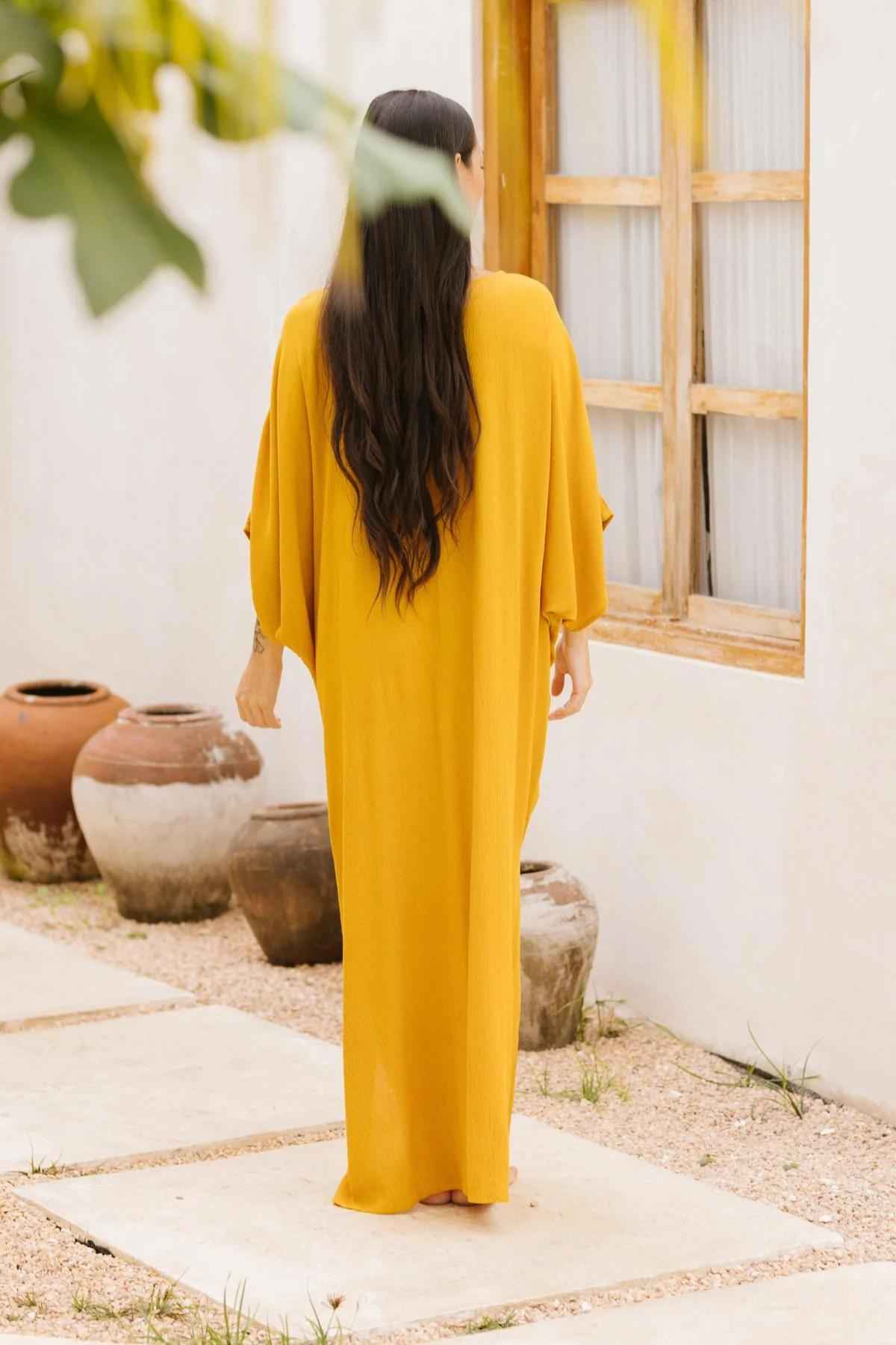 A woman with long, dark hair stands with her back to the camera, adorned in a sophisticated mustard-yellow dress that looks reminiscent of Myrah Penaloza's Limited Edition Virgo Kaftan. She is positioned near a white wall featuring a wooden-framed window. Several clay pots are scattered on the ground nearby, while green leaves partially frame the top of the image.