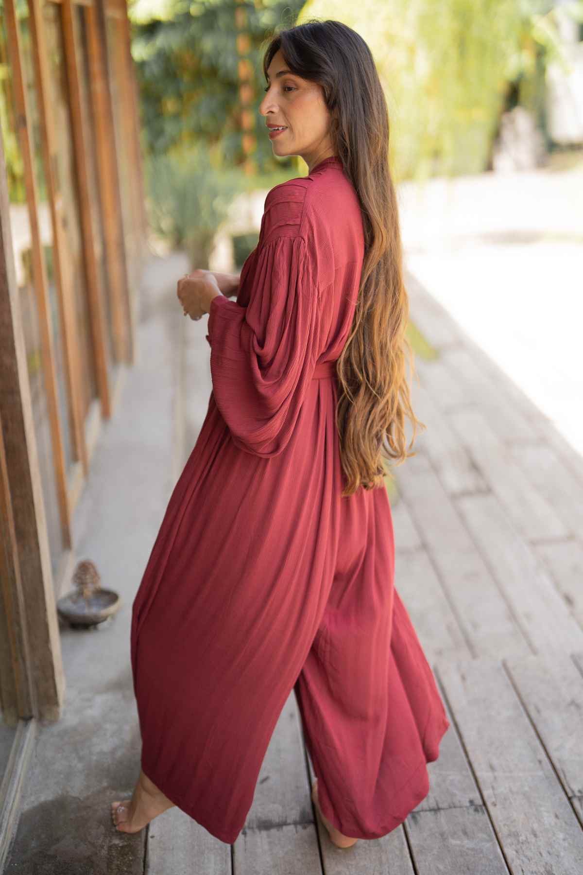 A person with long hair walks barefoot along a wooden pathway, adorned in the flowing, long-sleeved, red Kundalini Gown Playsuit Edition by Myrah Penaloza. The tranquil outdoor setting features trees and sunlight in the background, creating a magical ambiance perfect for an enchanting date night amidst nature's beauty.