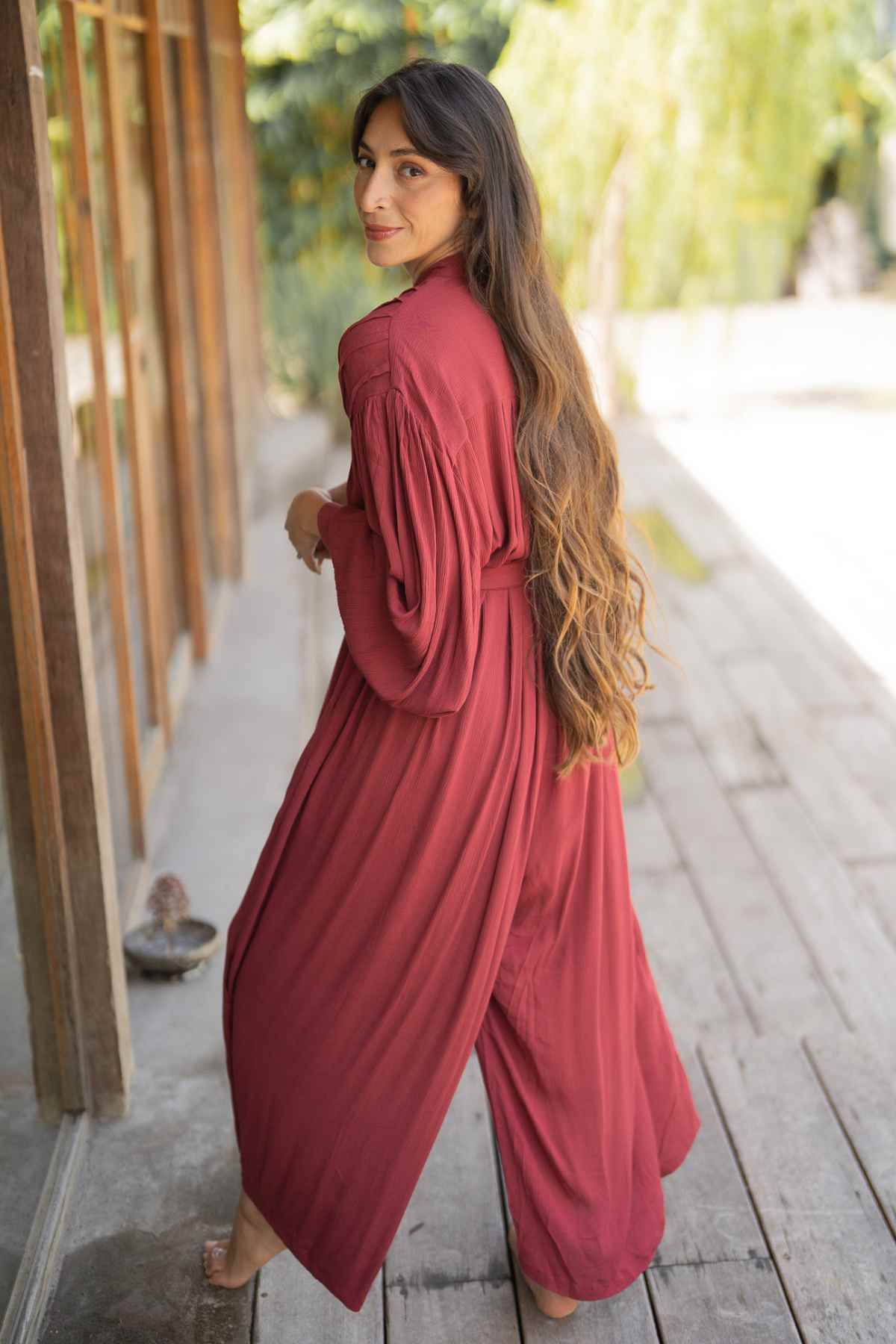 A person with long, wavy hair wears a flowing Kundalini Gown Playsuit Edition from Myrah Penaloza, featuring wide sleeves and a waist tie. They stand on a wooden path next to a wooden building, looking over their shoulder at the camera with a slight smile, surrounded by greenery.