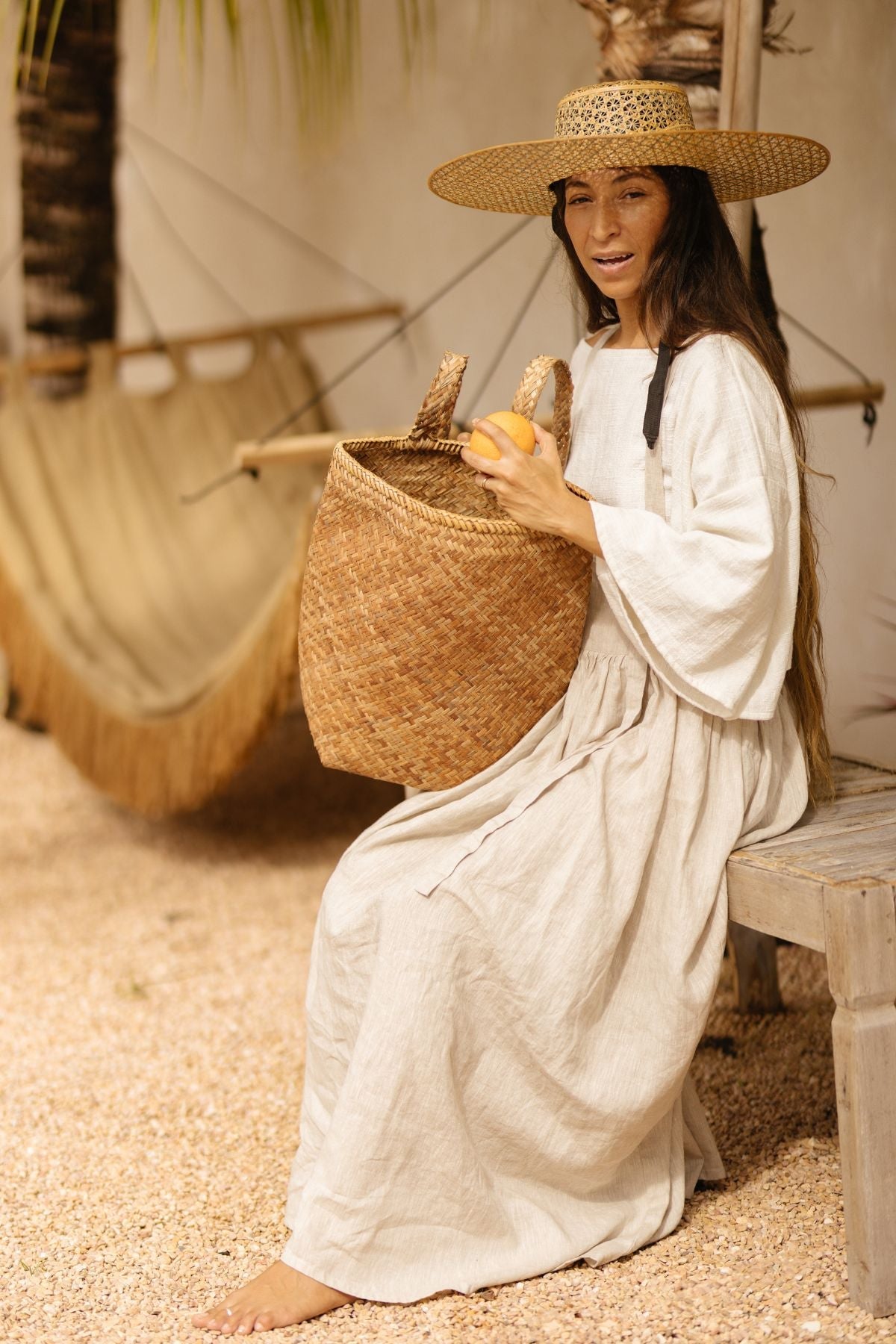 A woman clad in the May May Overall Skirt, a made-to-order piece by Myrah Penaloza, and a wide-brimmed straw hat, sits on a wooden bench. She holds a woven basket with a round yellow fruit inside. A hammock and palm tree are in the background, suggesting a relaxed, tropical setting.