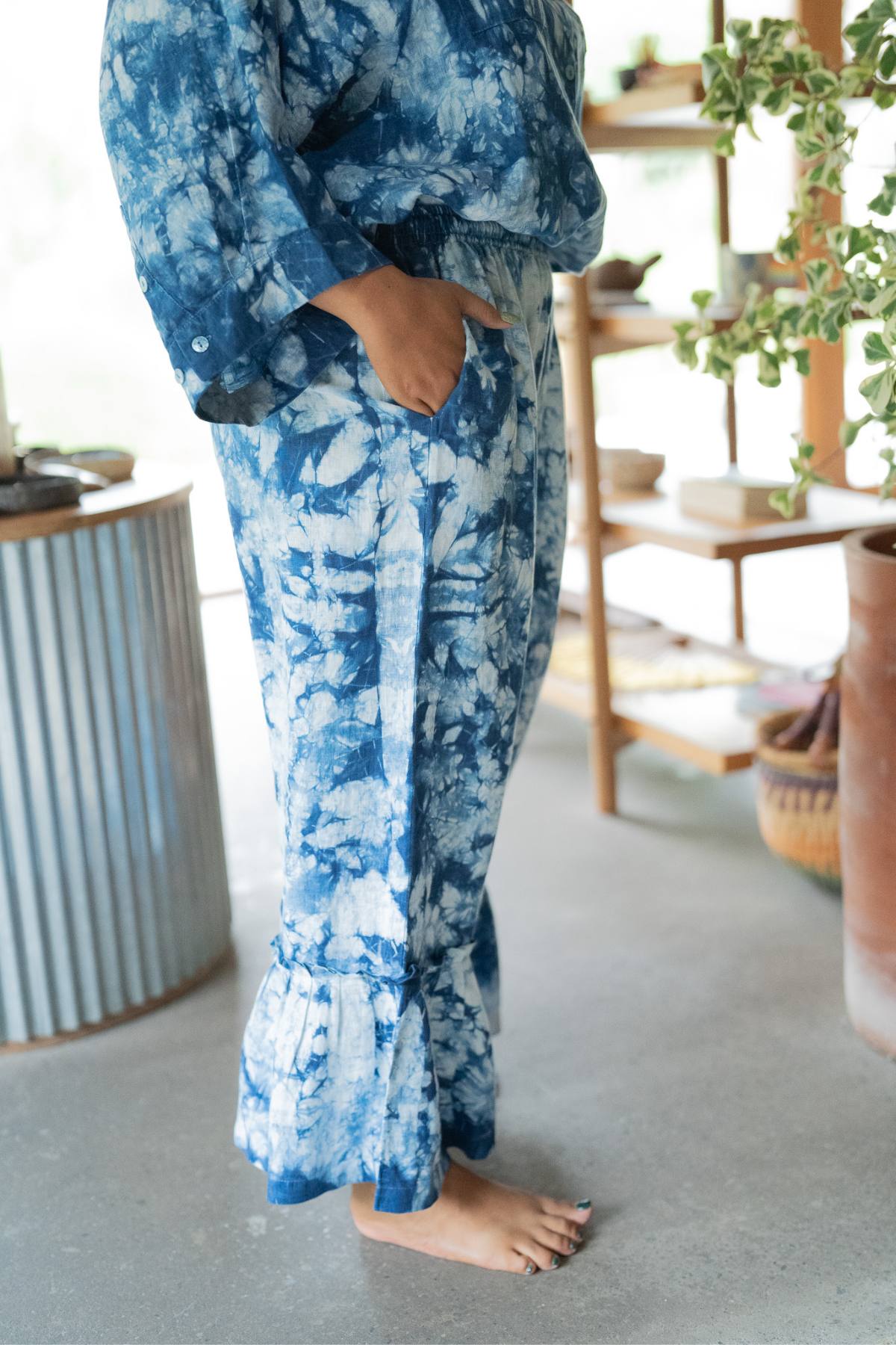 Barefoot and poised in a comfortable ensemble, the person wears blue tie-dye pants with ruffled cuffs and a matching top, reminiscent of Indigo Donna Pants by Myrah Penaloza. Shelves adorned with pottery and a potted plant offer dancing inspiration in the serene background.