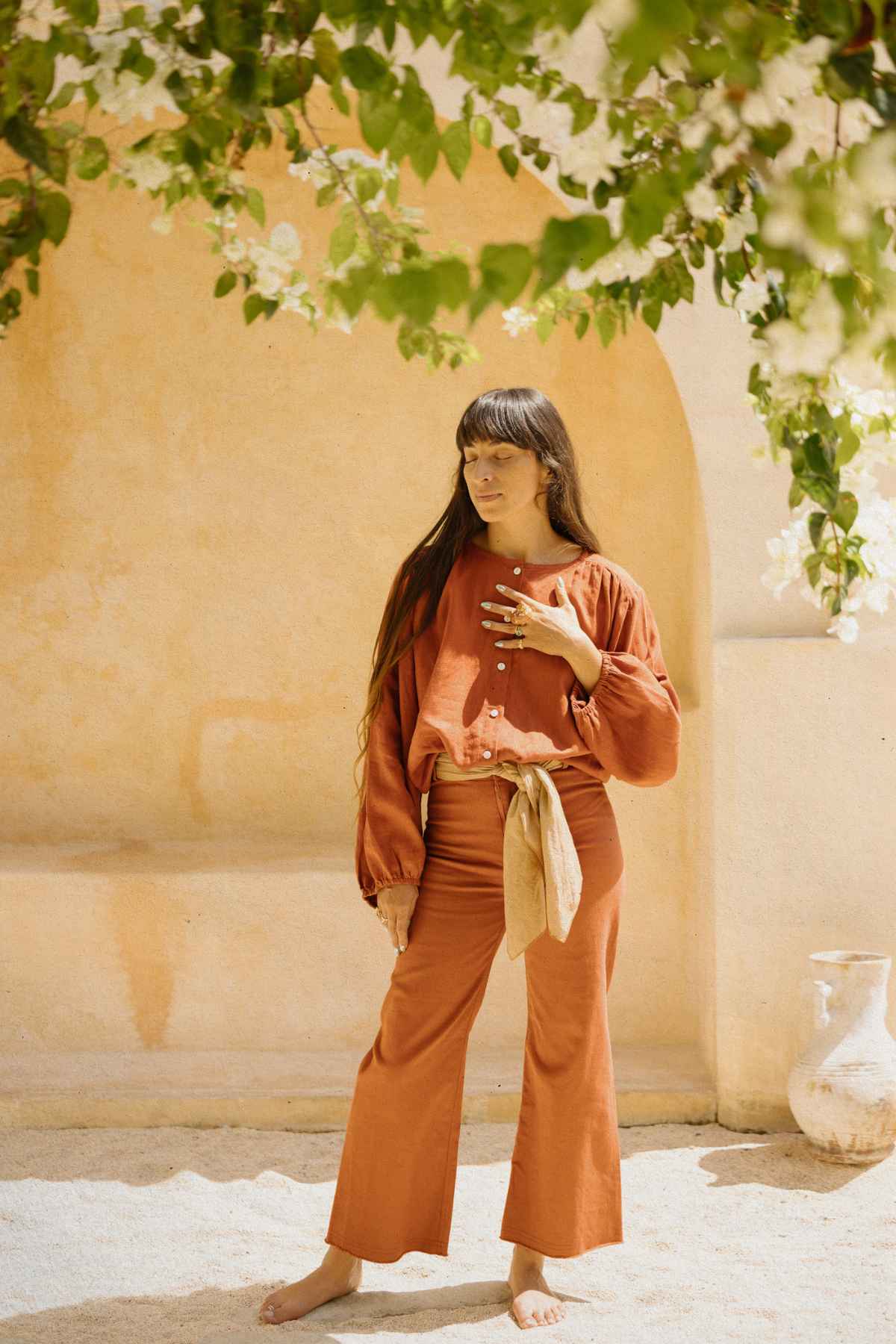 A woman with long hair stands barefoot on a sandy surface. She is wearing a botanically hand-dyed, stonewashed Vida high-waisted jean by Myrah Penaloza, paired with an orange naturally hand-dyed long-sleeve shirt and a beige cloth belt. She touches her collar with one hand and looks away, with a leafy plant overhead and an archway in the background.