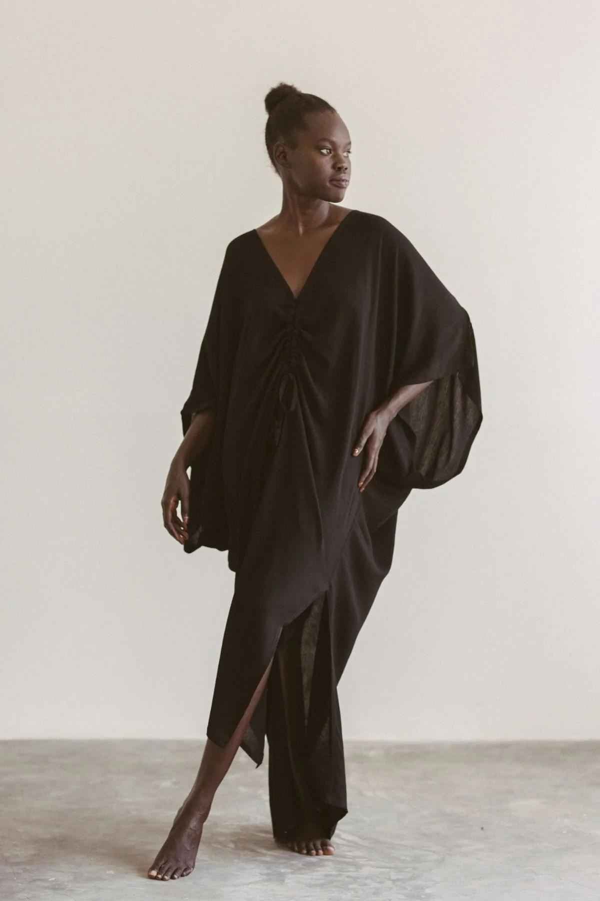 A person stands barefoot on a neutral-colored floor, wearing the sophisticated Limited Edition Virgo Kaftan in black by Myrah Penaloza, which features a deep V-neck. The flowing outfit, made from luxe bamboo rayon, includes draped sleeves and gathered detail at the waist. They look to their left with a neutral expression against a plain white wall.