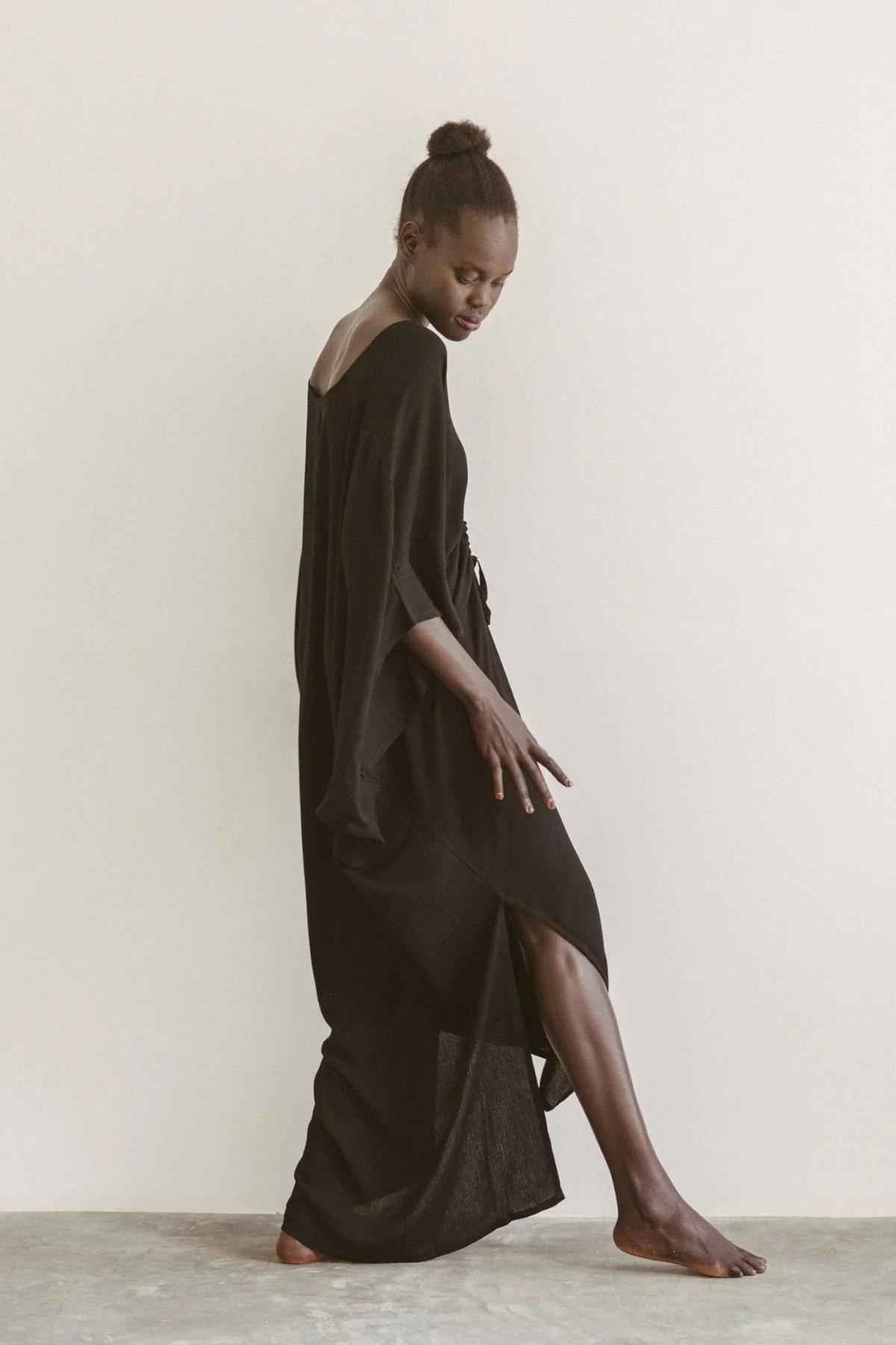 A person with dark skin and a high bun hairstyle poses gracefully against a plain backdrop. They are wearing the Limited Edition Virgo Kaftan by Myrah Penaloza, a long, flowing black dress with a slit that reveals part of their leg. One arm is slightly extended, and their head is turned downward in sophisticated fashion.