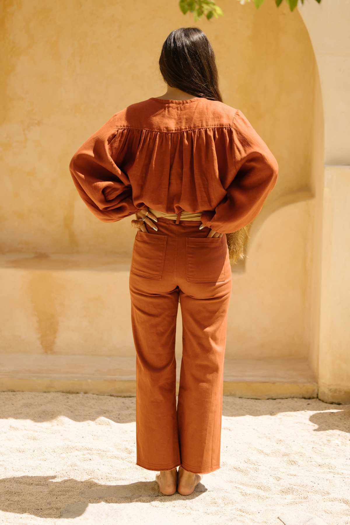 A person with long hair stands with their back to the camera, wearing a matching autumnal brown outfit from Myrah Penaloza's Rice Linen Set, which includes wide-leg pants and an elegant stone-washed top featuring voluminous sleeves. Their hands are resting on their lower back as they stand barefoot on a sandy surface.