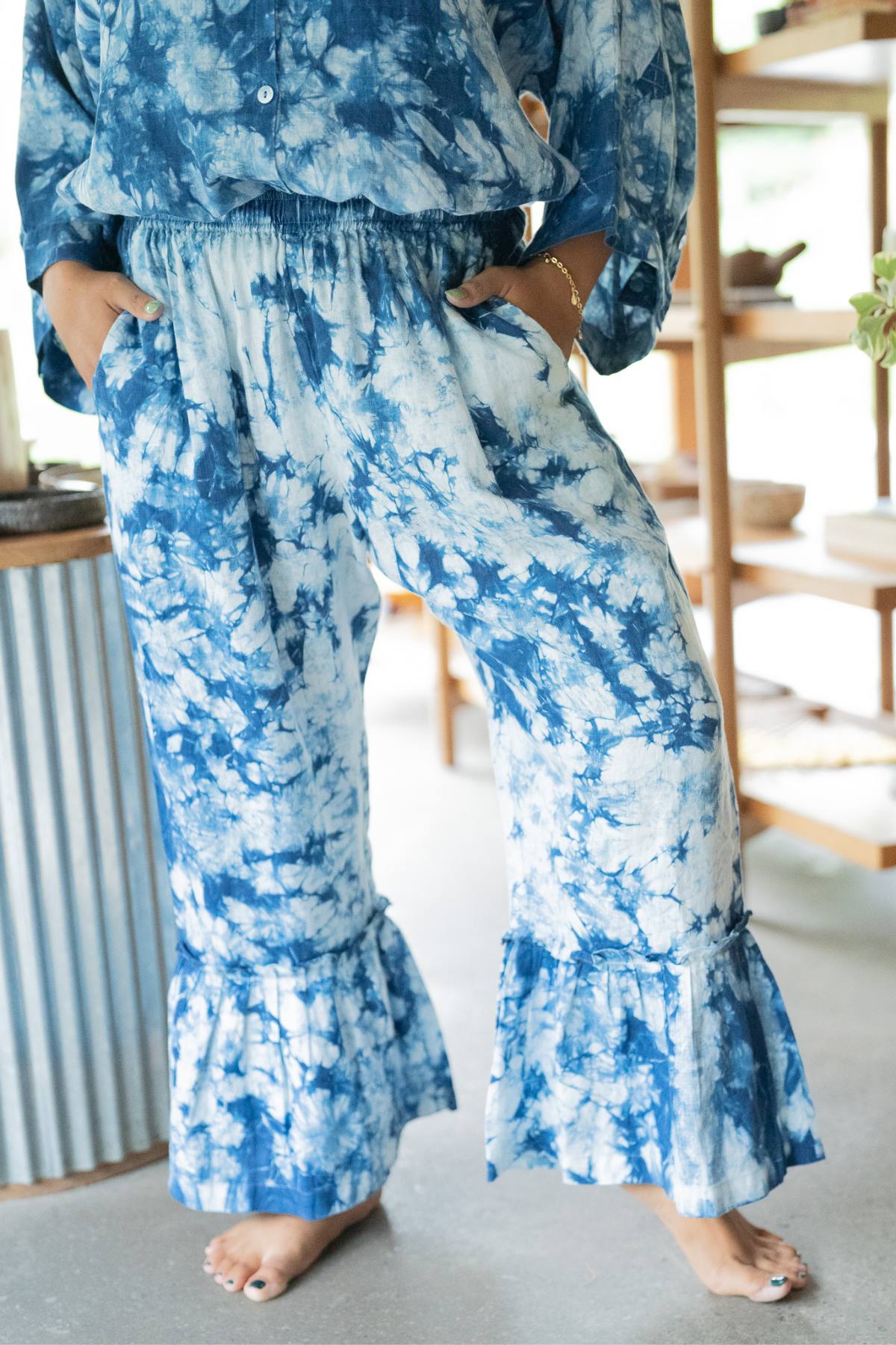 A person wearing Indigo Donna Pants by Myrah Penaloza, in a unique blue and white tie-dye pattern with ruffled hems, stands effortlessly with hands in pockets, exuding a dance-inspiring flow and comfort.