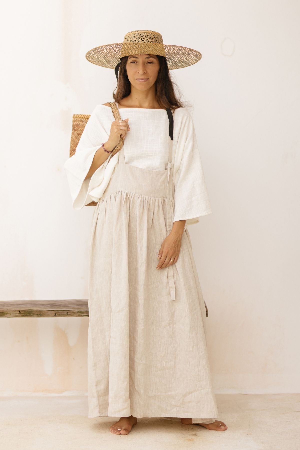 A person with long hair stands against a plain background, wearing a wide-brimmed straw hat, a white long-sleeve blouse, and the beige May May Overall Skirt by Myrah Penaloza. They hold a woven bag on their shoulder and have a calm expression.