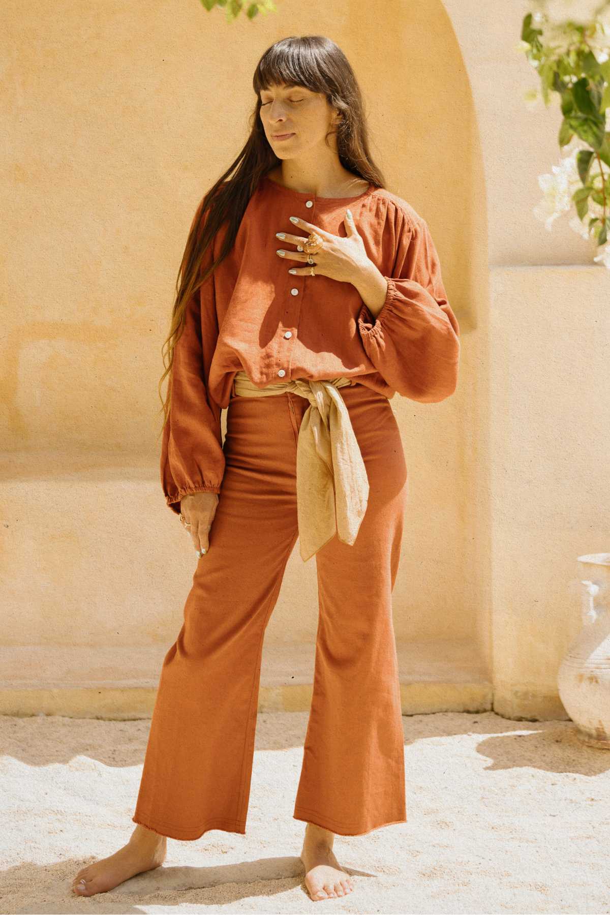 A woman stands barefoot on light-colored sand with a beige background, elegantly dressed in the Rice Linen Set by Myrah Penaloza. The outfit features an orange long-sleeve top with white buttons and matching high-waisted pants. A cream-colored scarf is gracefully draped around her waist, and she has long dark hair and bangs, showcasing versatile fashion.