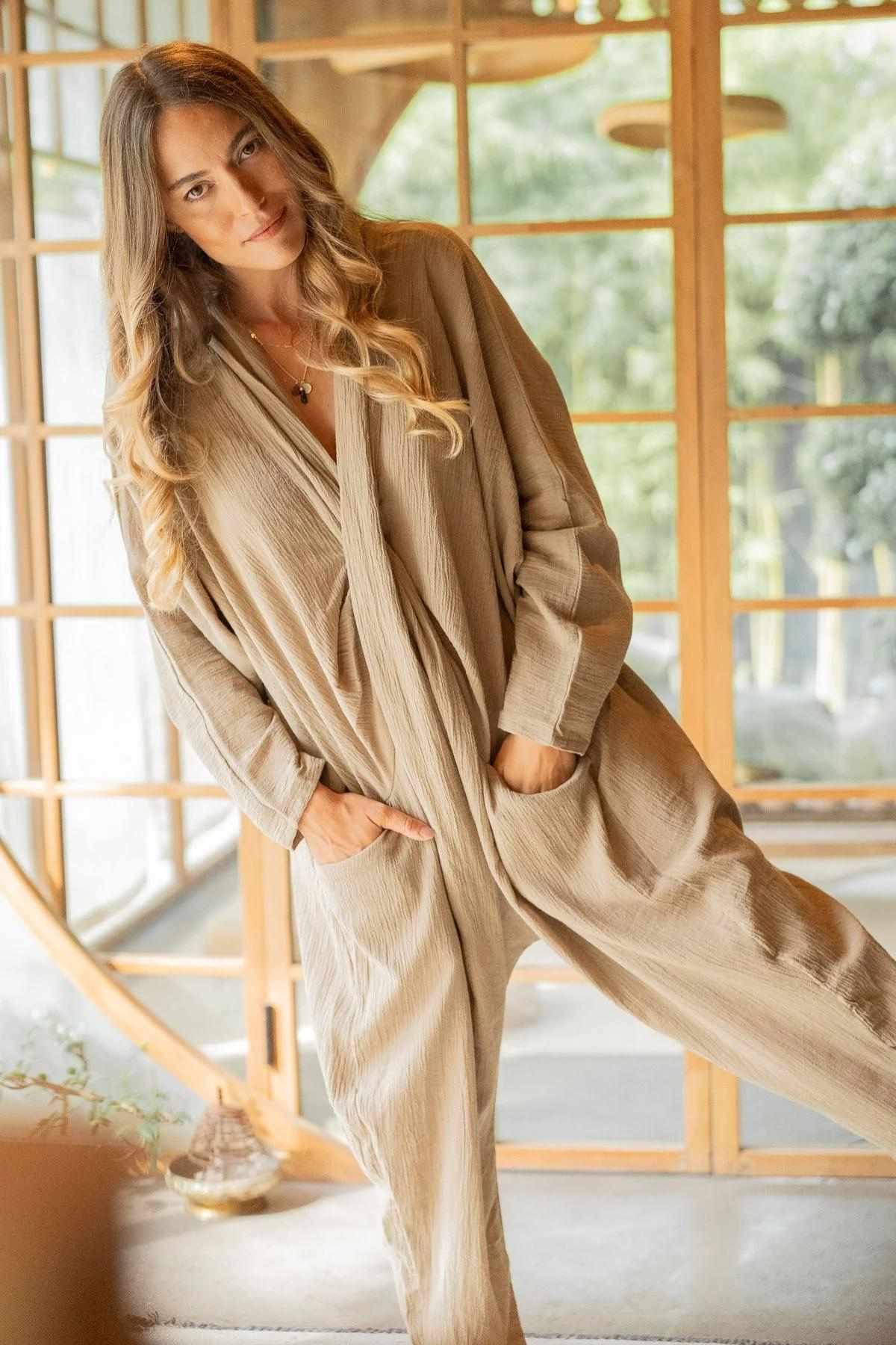 A woman with long, wavy hair is posing indoors near a wooden-framed window. She is wearing a Myrah Penaloza Crinkle Linen Amma Playsuit with her hands in the pockets. The background shows greenery outside, creating a serene atmosphere.