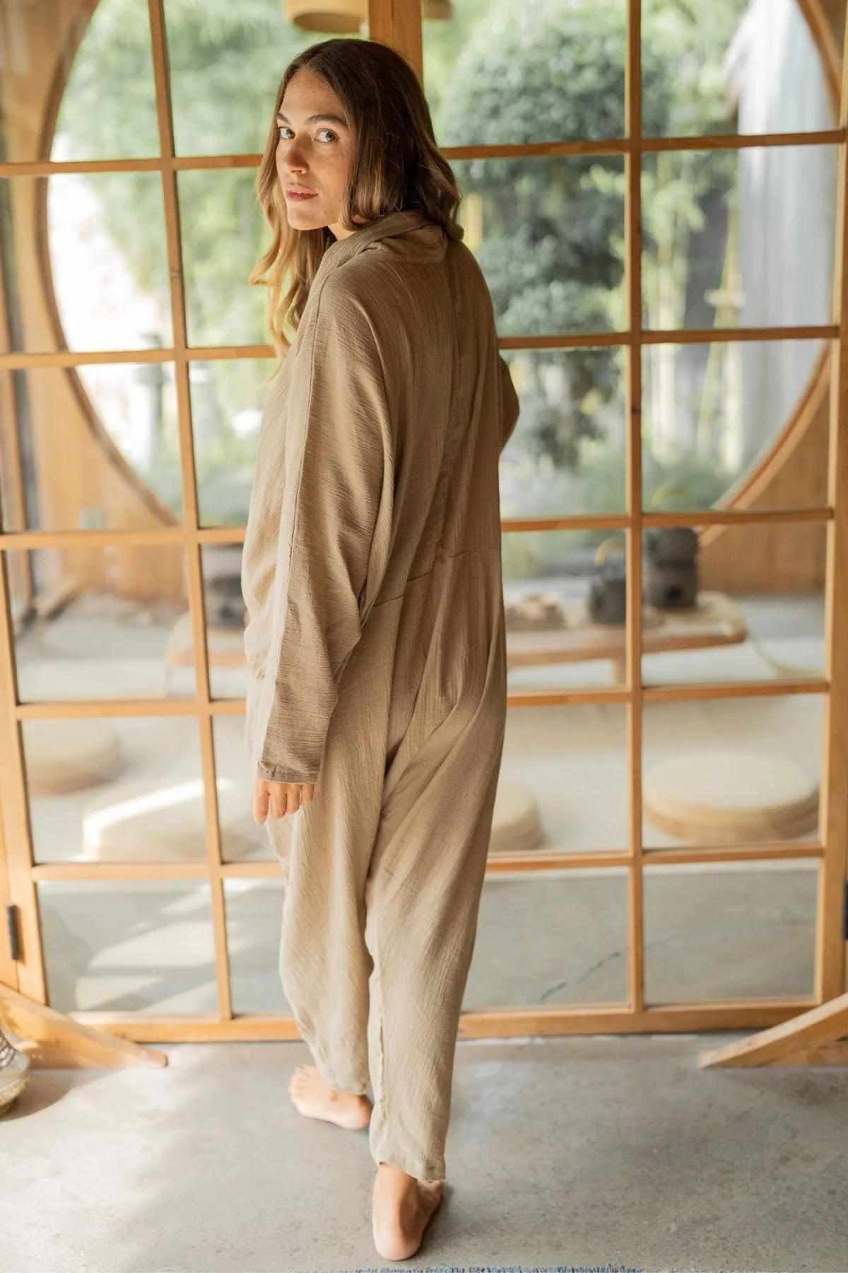 A person stands barefoot on a stone floor, wearing an oversized Crinkle Linen Amma Playsuit from Myrah Penaloza, and looking over their shoulder. The background features wooden framed glass doors, revealing greenery outside and a serene indoor setting.