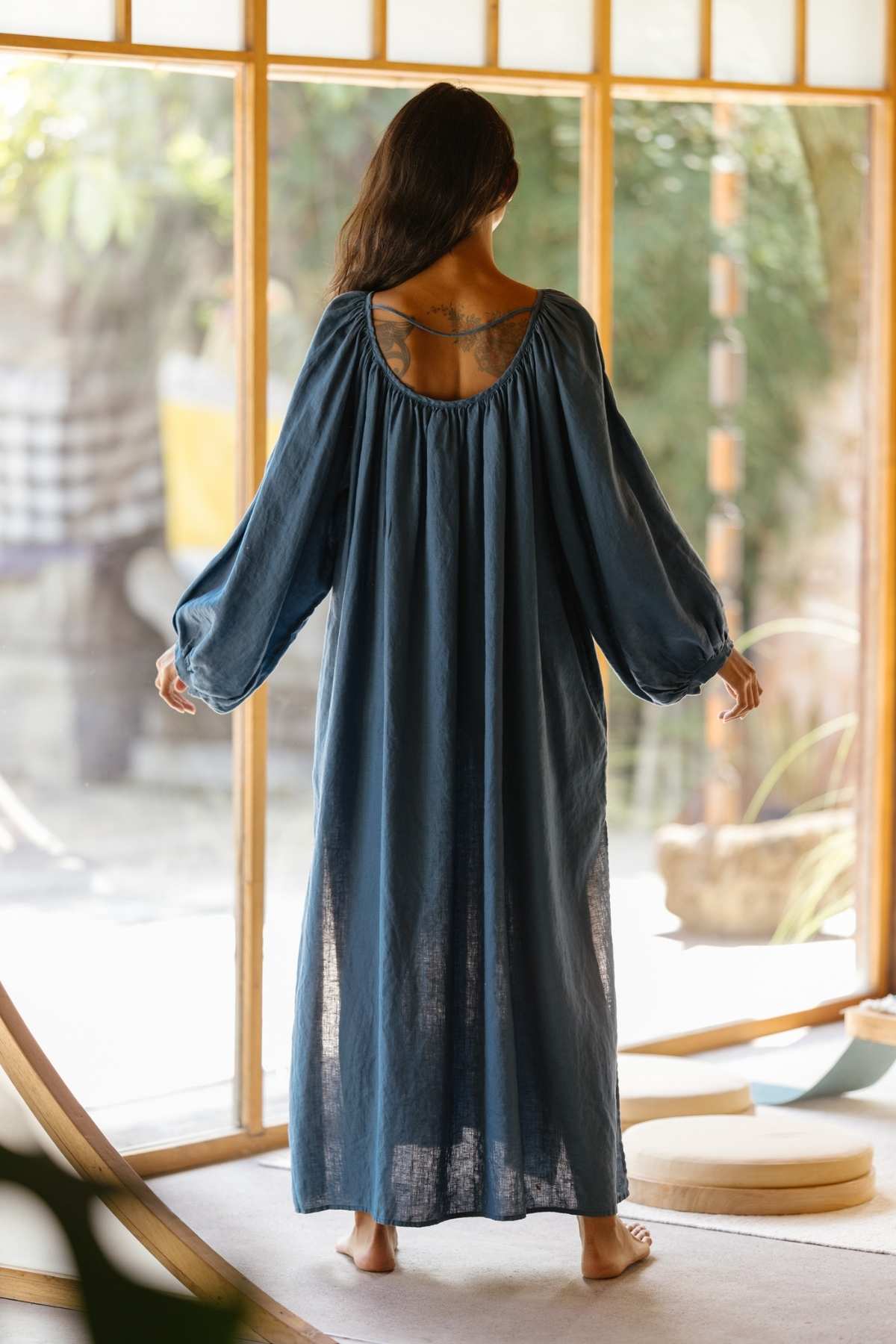 A woman stands barefoot in front of a large window illuminated by natural light, adorned in a long, flowing blue Alchemy Gown (Crinkle Linen Cotton) by Myrah Penaloza, featuring elegant puffy sleeves. With her back to the camera, she reveals a tattoo on her upper back. The serene room boasts a wooden floor and minimal decor, creating an ambiance reminiscent of a modern mystic.